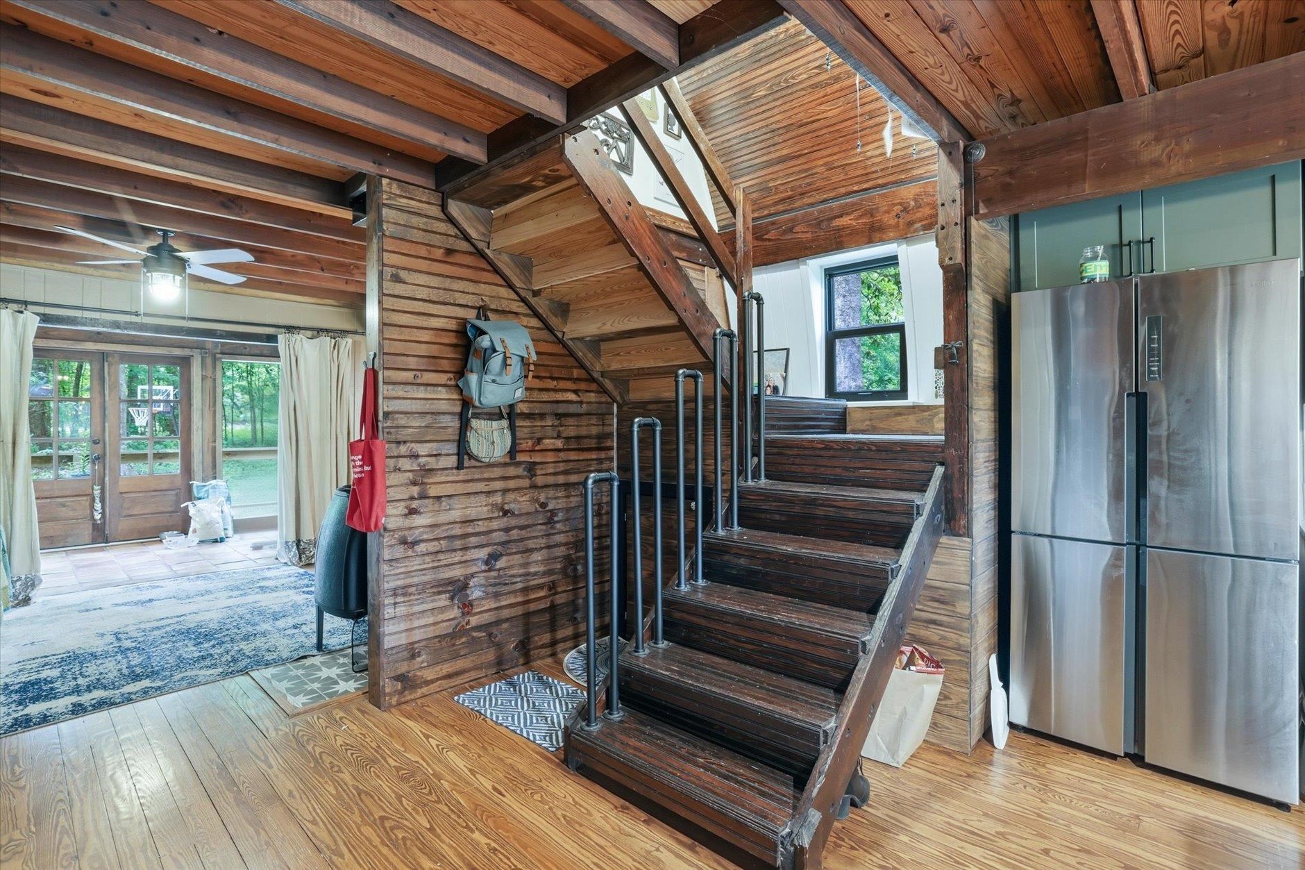 Wooden staircase with metal railings beneath exposed beams, beside a stainless-steel refrigerator.