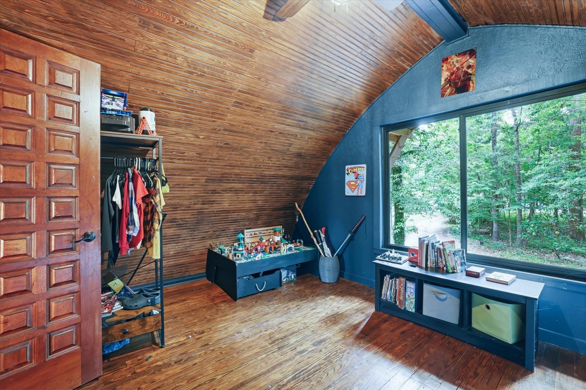 Attic playroom with a sloped wood ceiling, toys, and a large window overlooking trees.