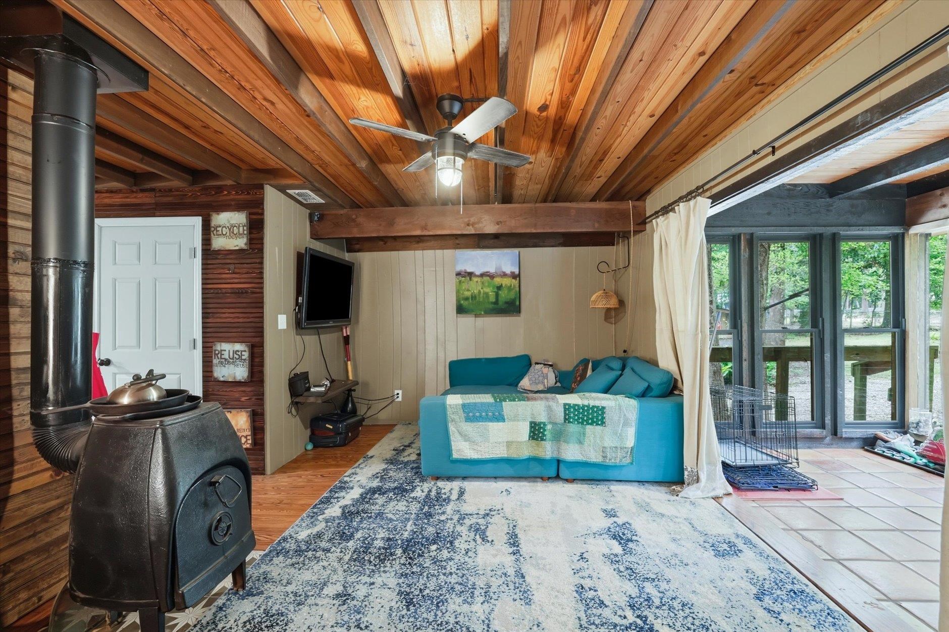 Living room with a wood-plank ceiling, black wood stove, turquoise sectional, and large windows overlooking trees.