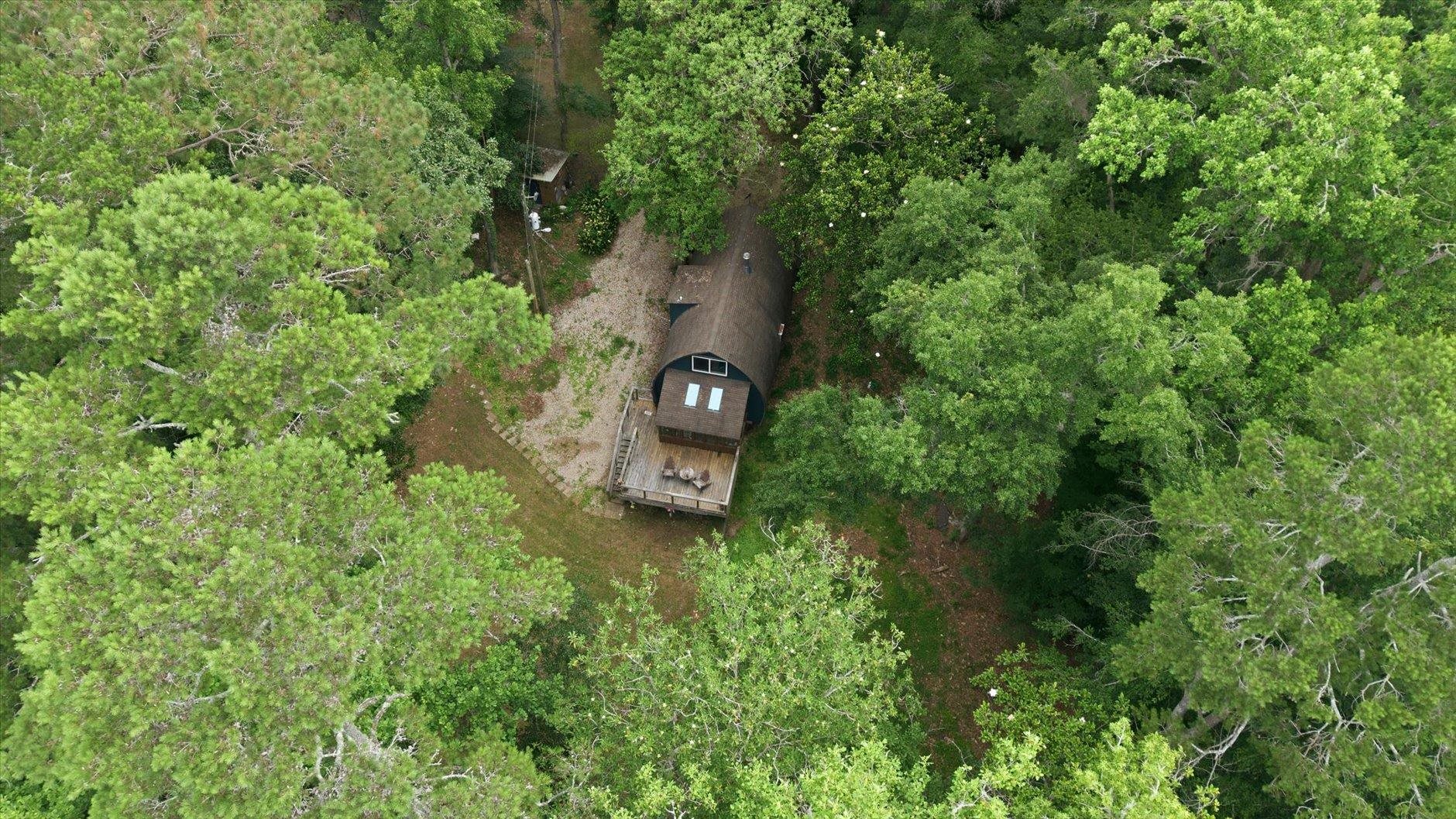 Aerial view of a cabin and wooden deck nestled among dense trees beside a gravel driveway.
