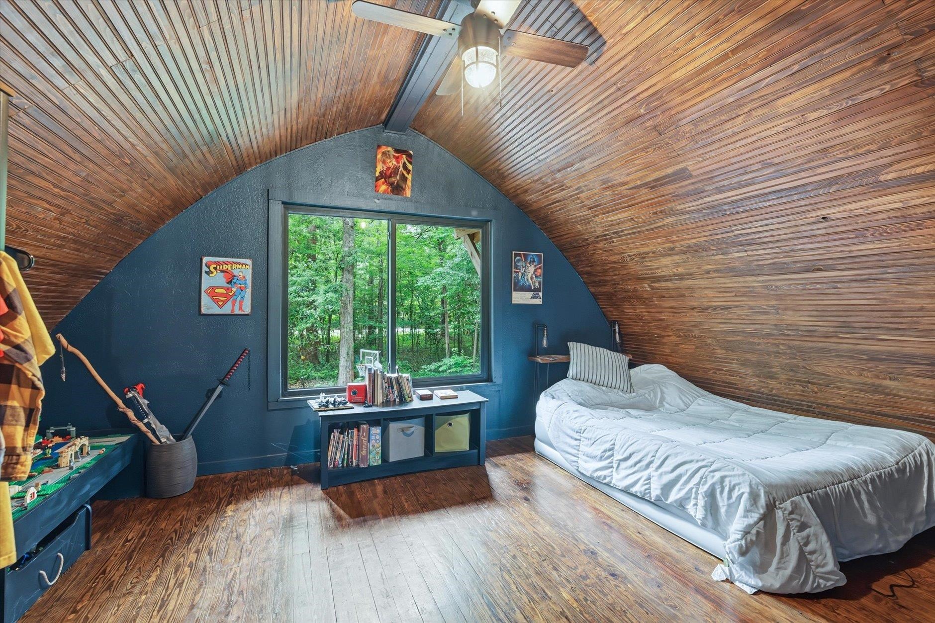 Attic bedroom with a vaulted wood ceiling, dark blue walls, and a large window overlooking trees.