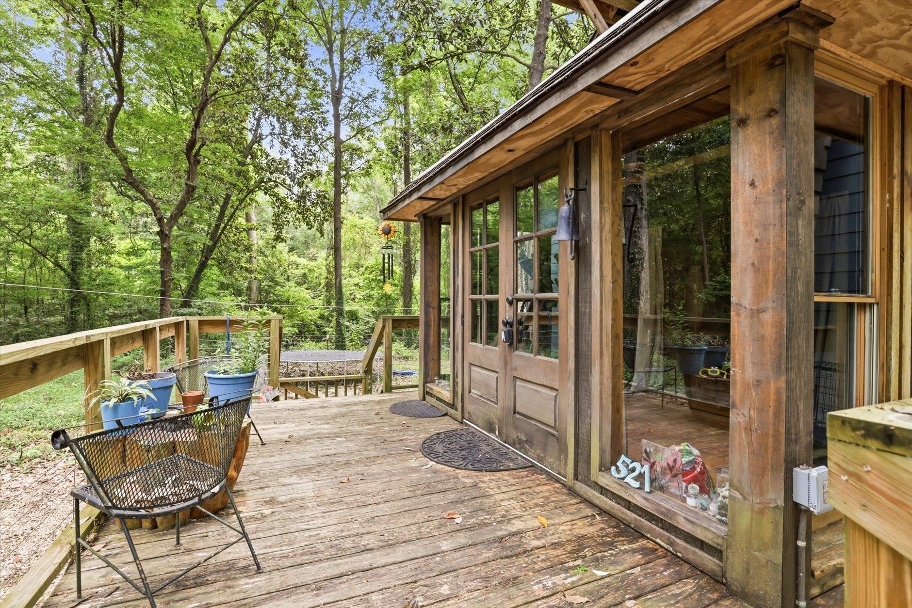 Wooden deck beside a rustic home, with glass-paneled doors and potted plants among leafy trees.