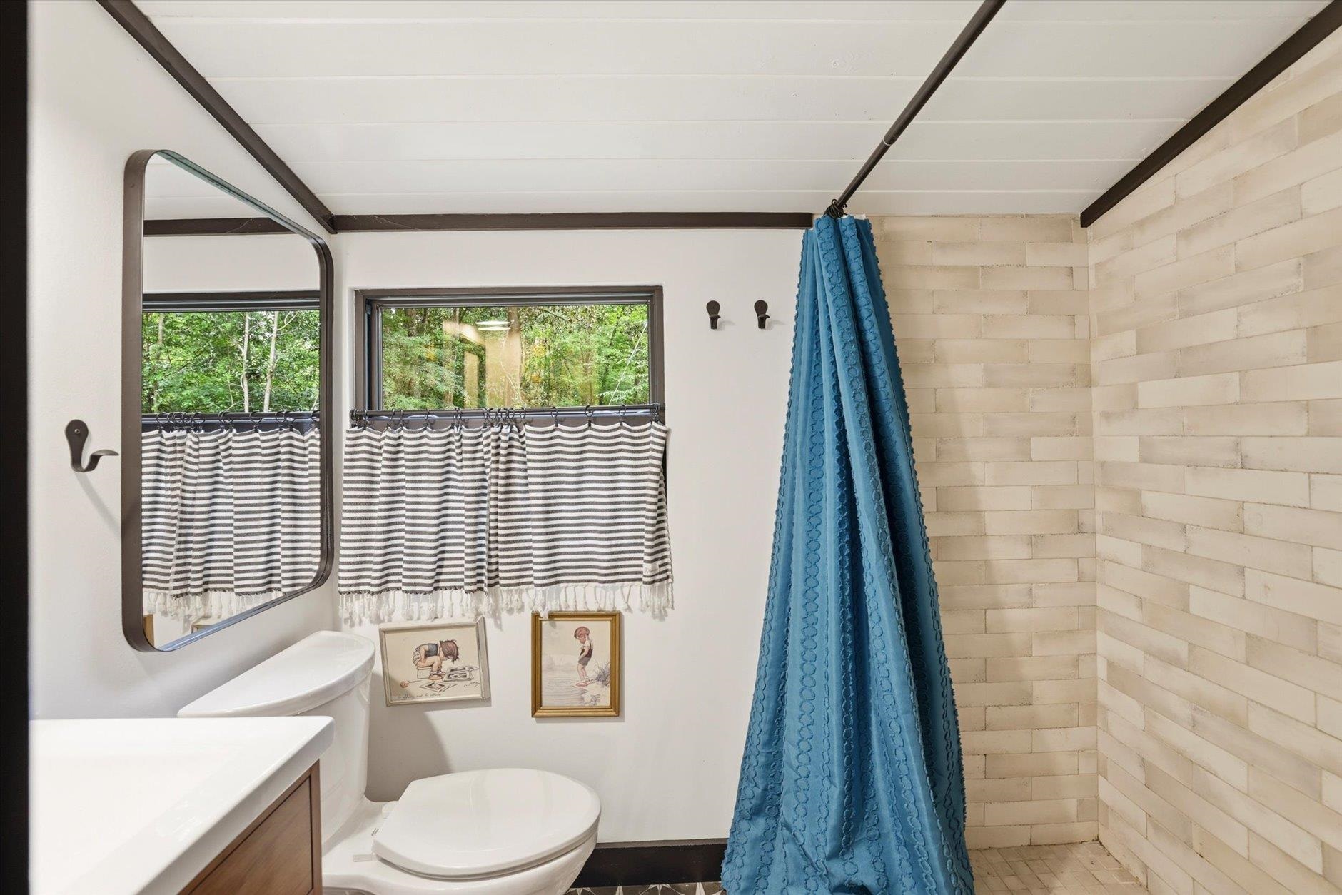 Bathroom with pale tiled shower, blue curtain, wood-front vanity, and window overlooking trees.