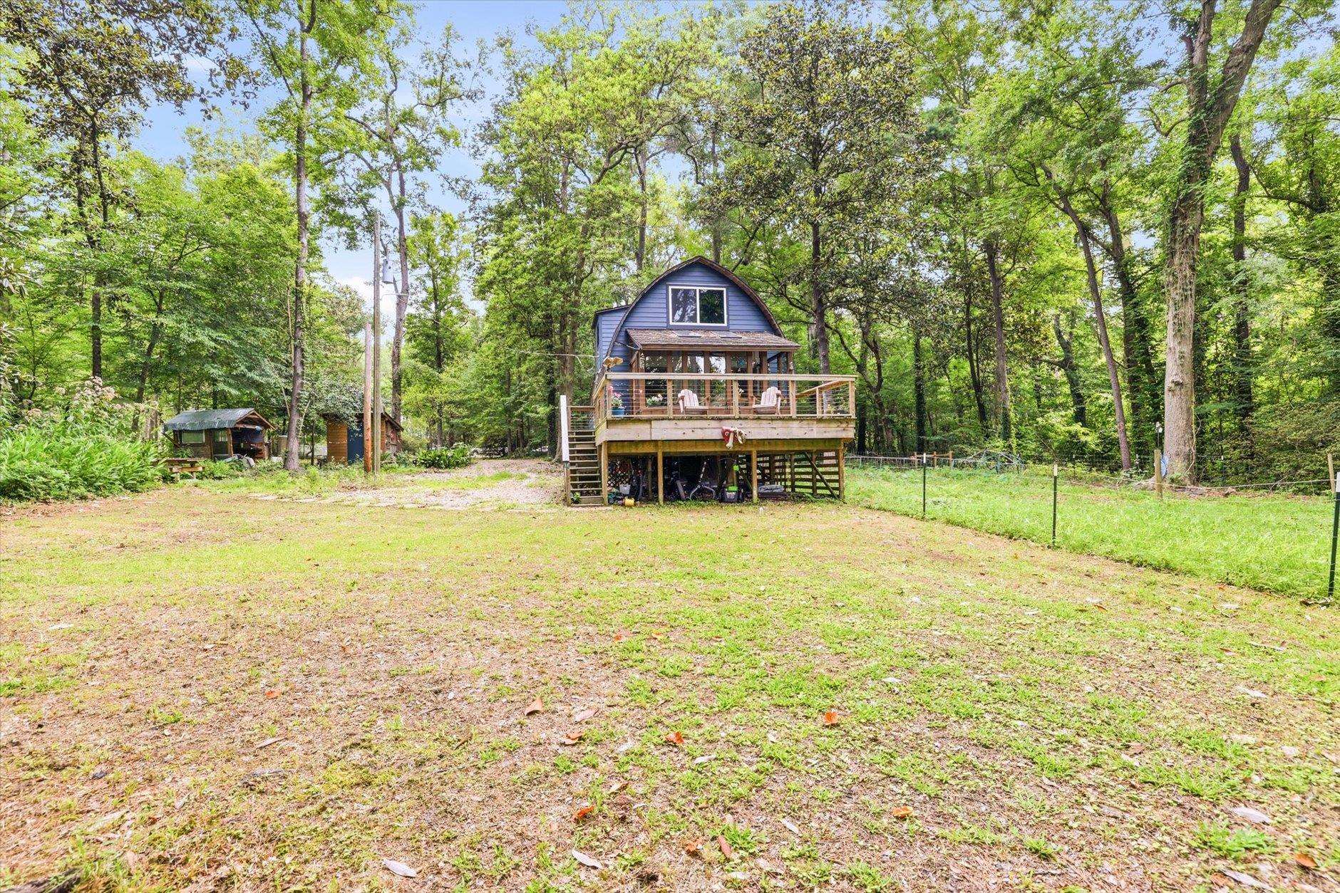 Blue house with a raised deck in a wooded clearing, with a small outbuilding nearby.