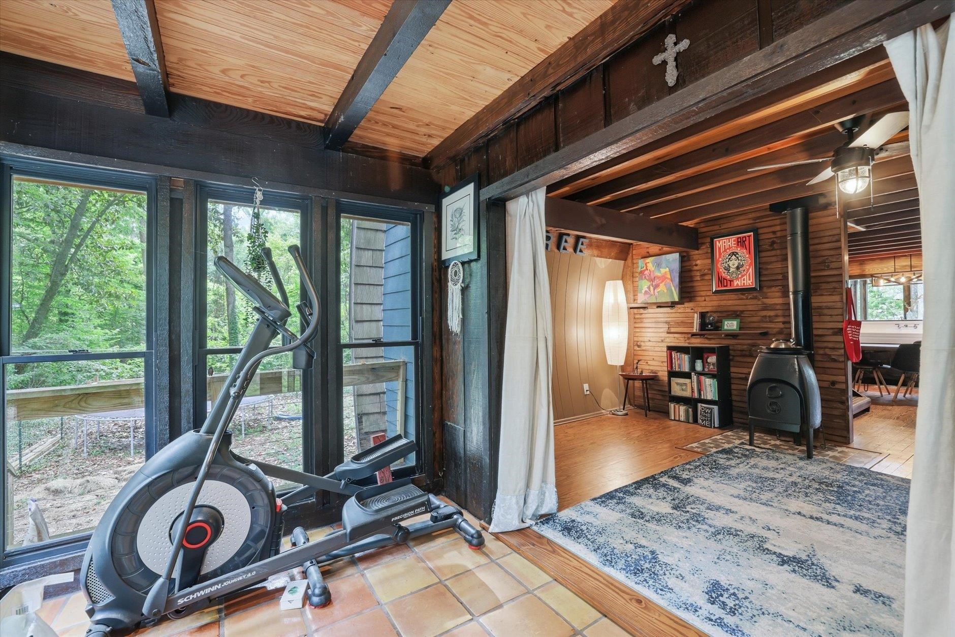 Rustic exercise room with an elliptical, large windows, exposed wood beams, and an adjoining wood-stove living area.