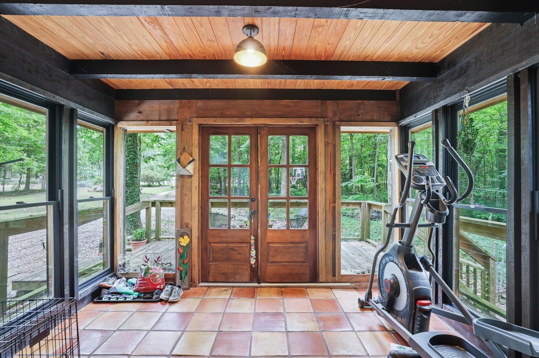 Enclosed sunroom with terracotta tile, wooden double doors, and an elliptical beside forest-facing windows.