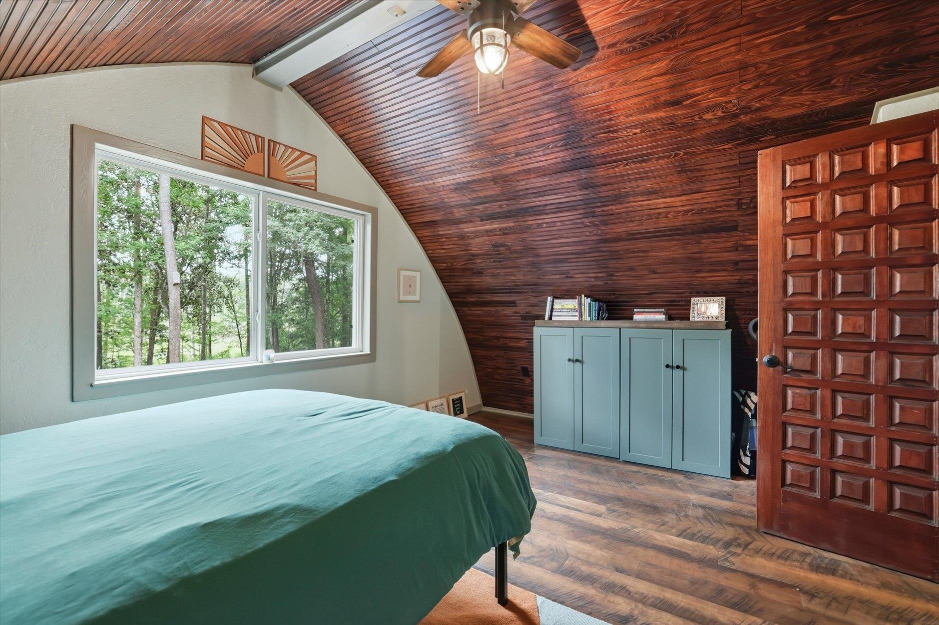 Bedroom with a dramatic wood-paneled vaulted ceiling, teal bedspread, and window overlooking trees.