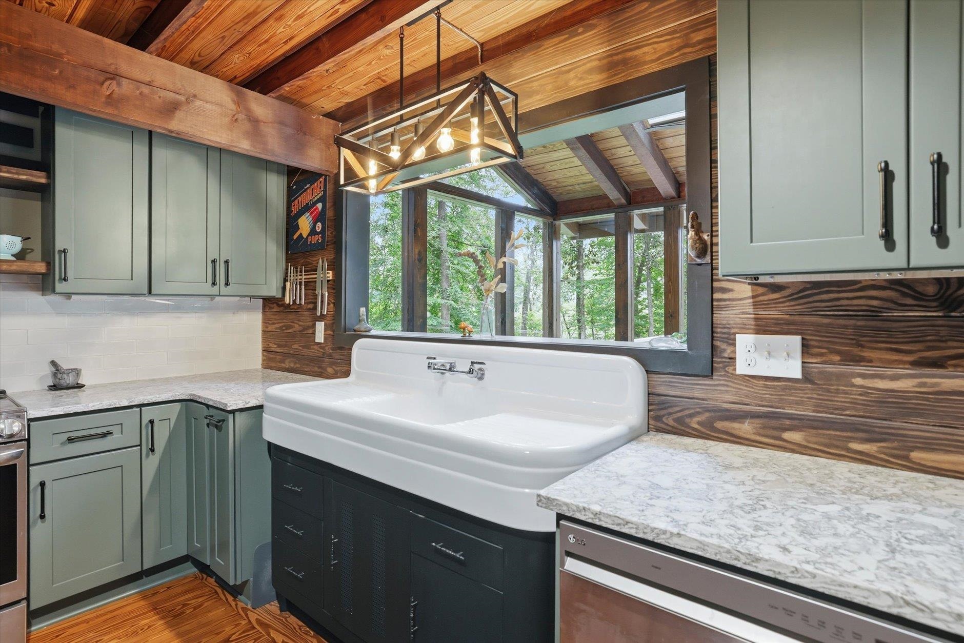 Kitchen with a farmhouse sink, sage-green cabinets, and vaulted wood ceiling overlooking trees.
