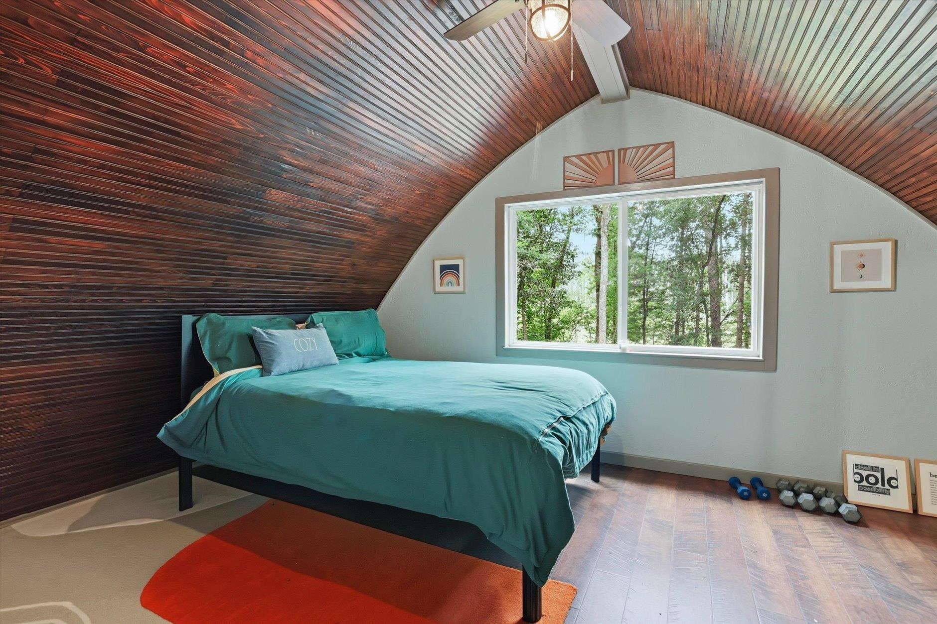 Bedroom with a teal bed beneath a dark wood-plank vaulted ceiling and a window overlooking trees.
