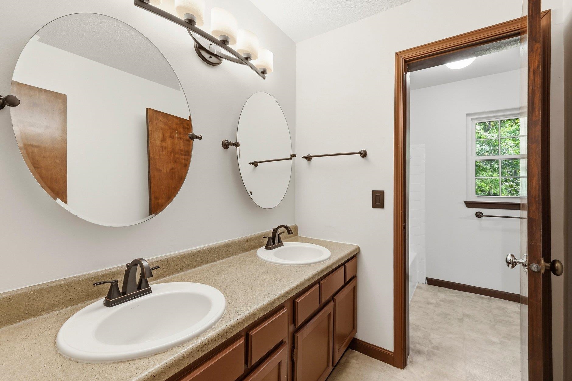 Bathroom with a double-sink vanity, oval mirrors, and wood cabinetry beside an open doorway.