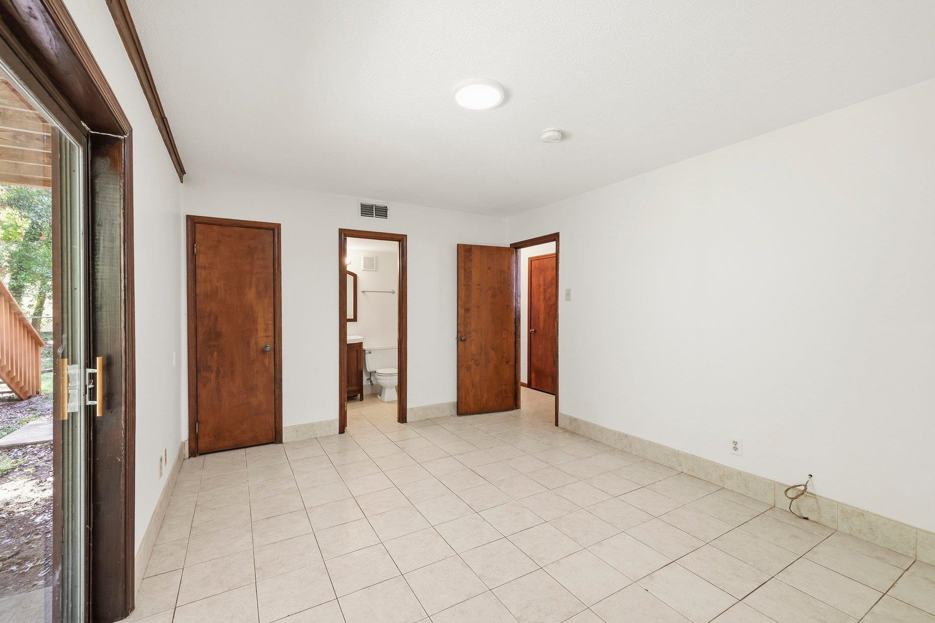 Vacant tiled room with dark wood doors, a bathroom doorway, and a sliding glass door to a deck.