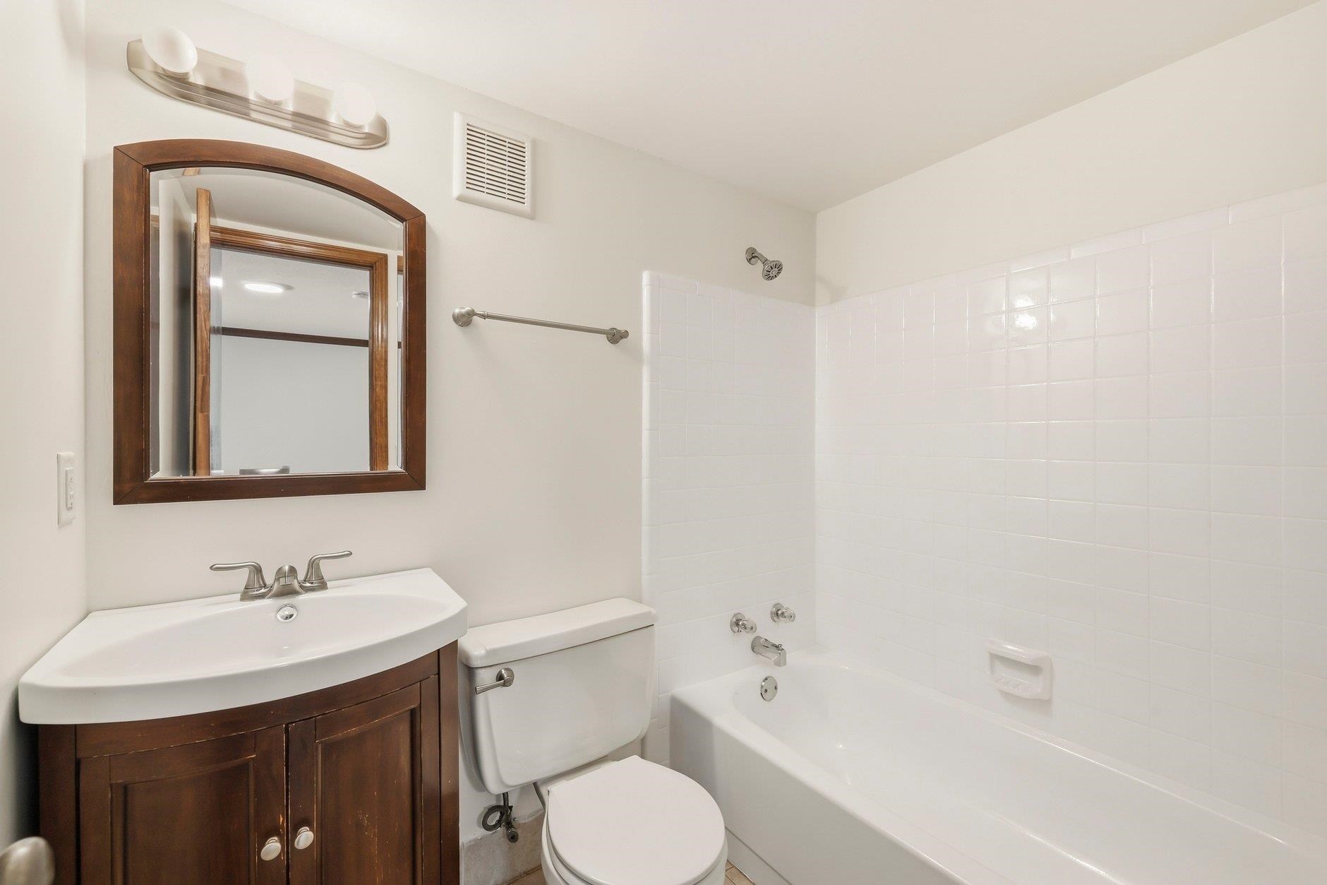 Bathroom with a white tiled tub and shower beside a dark wood vanity and arched mirror.