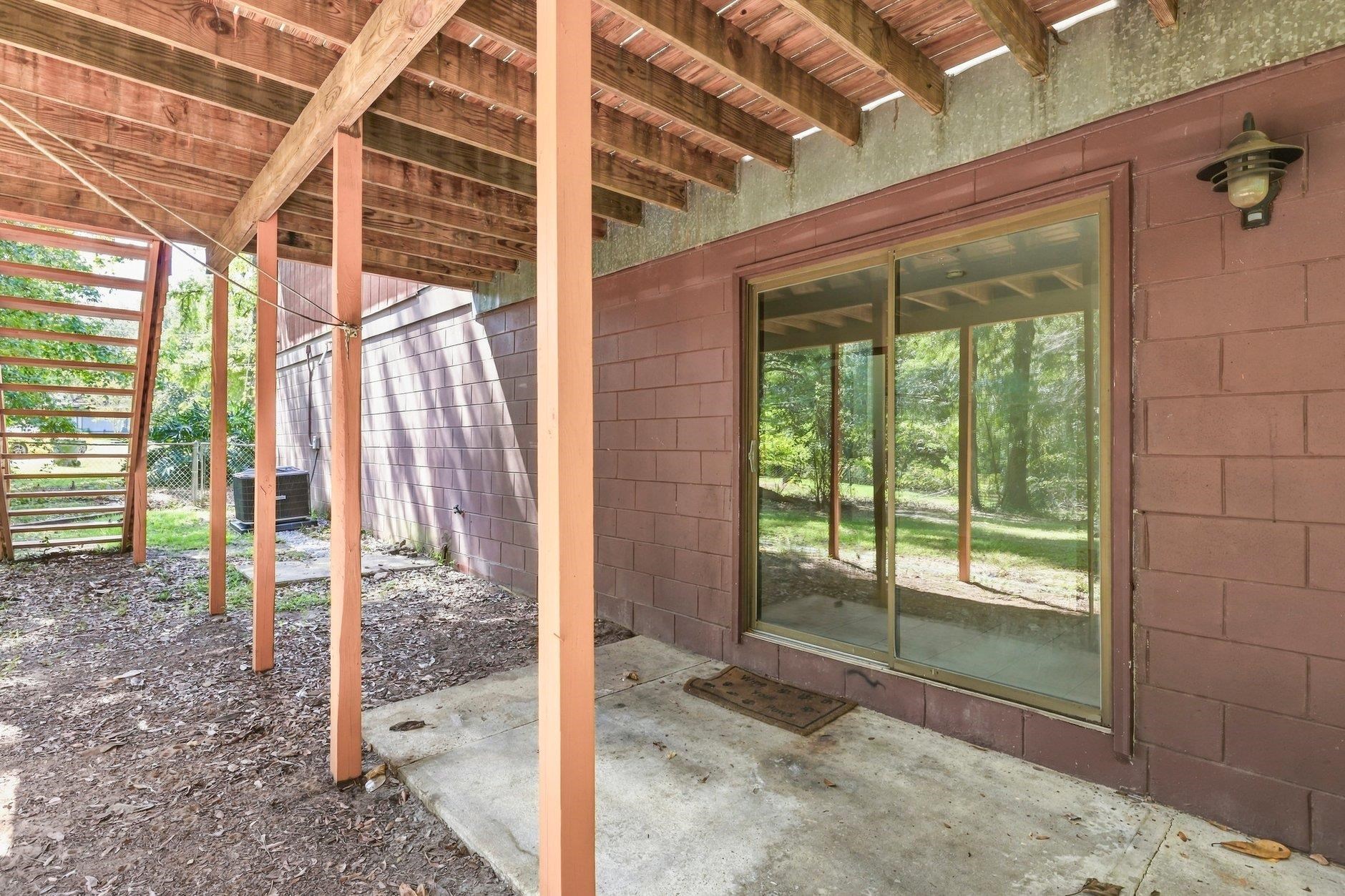 Covered patio with exposed wooden beams, concrete floor, and sliding glass doors overlooking a leafy yard.