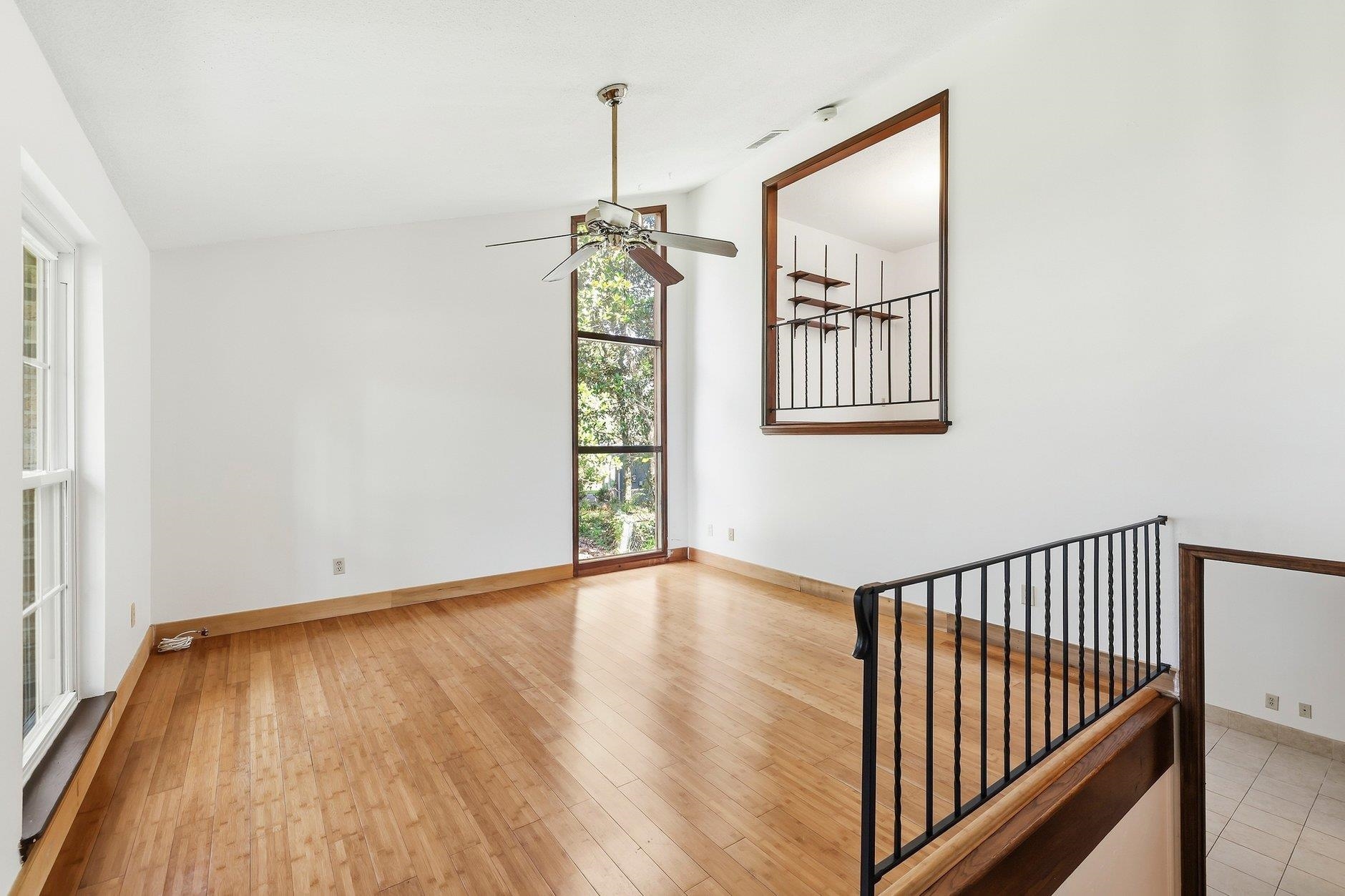 Empty hardwood-floored room with a ceiling fan, tall windows, and a railing around the stairwell.