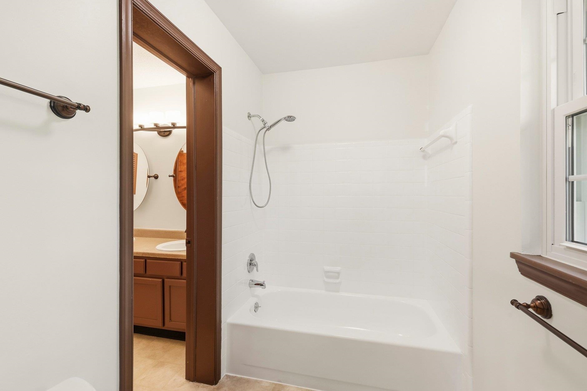 Bathroom with a white tub and tiled shower, beside an open doorway to a wood-tone vanity.