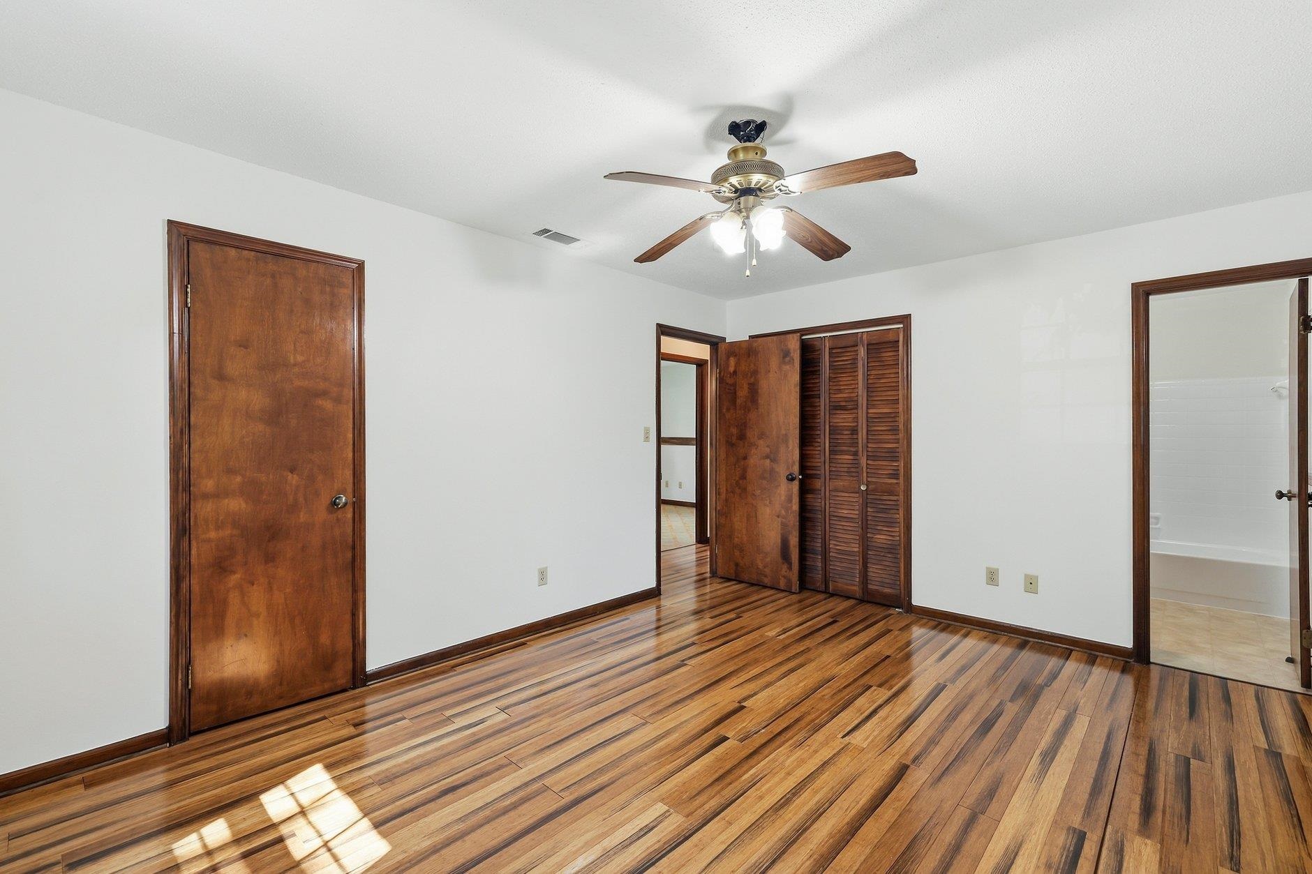 Empty bedroom with polished hardwood floors, dark wood doors, louvered closet doors, and a ceiling fan.