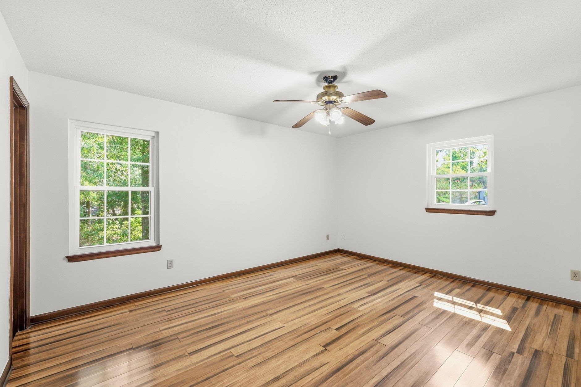 Empty room with polished hardwood floors, two windows, dark wood trim, and a ceiling fan.