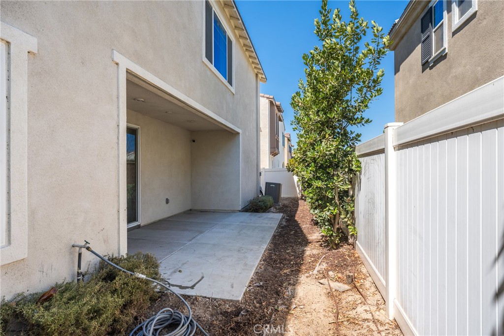 Side yard with a concrete patio, white fence, and mature tree beside a two-story stucco home.