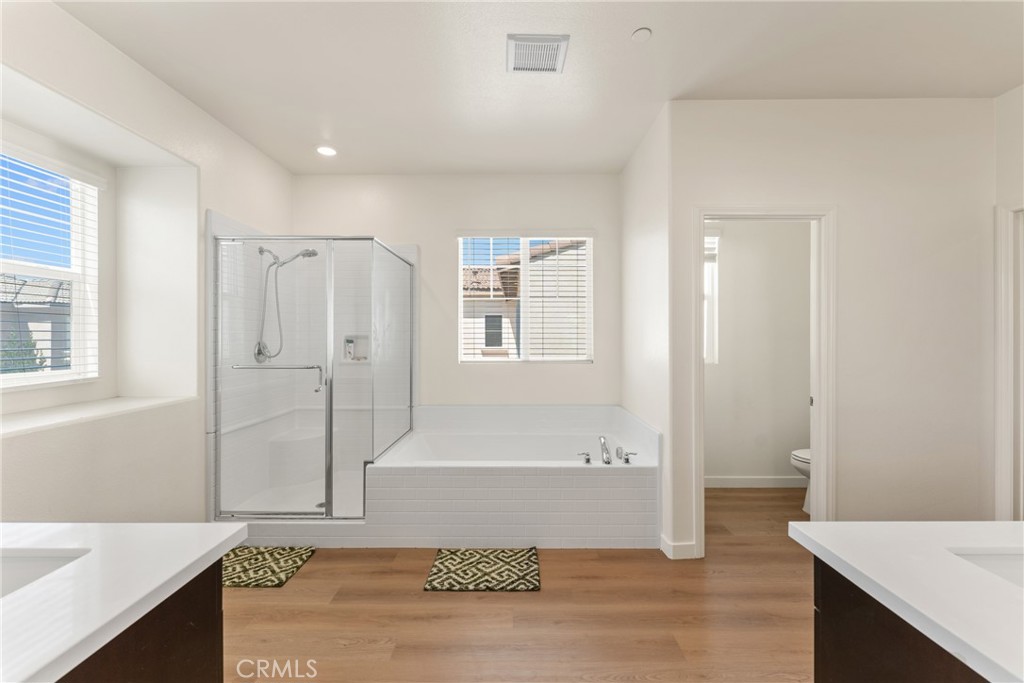 Bright bathroom with a glass shower, built-in tub, dual vanities, and wood-look flooring.