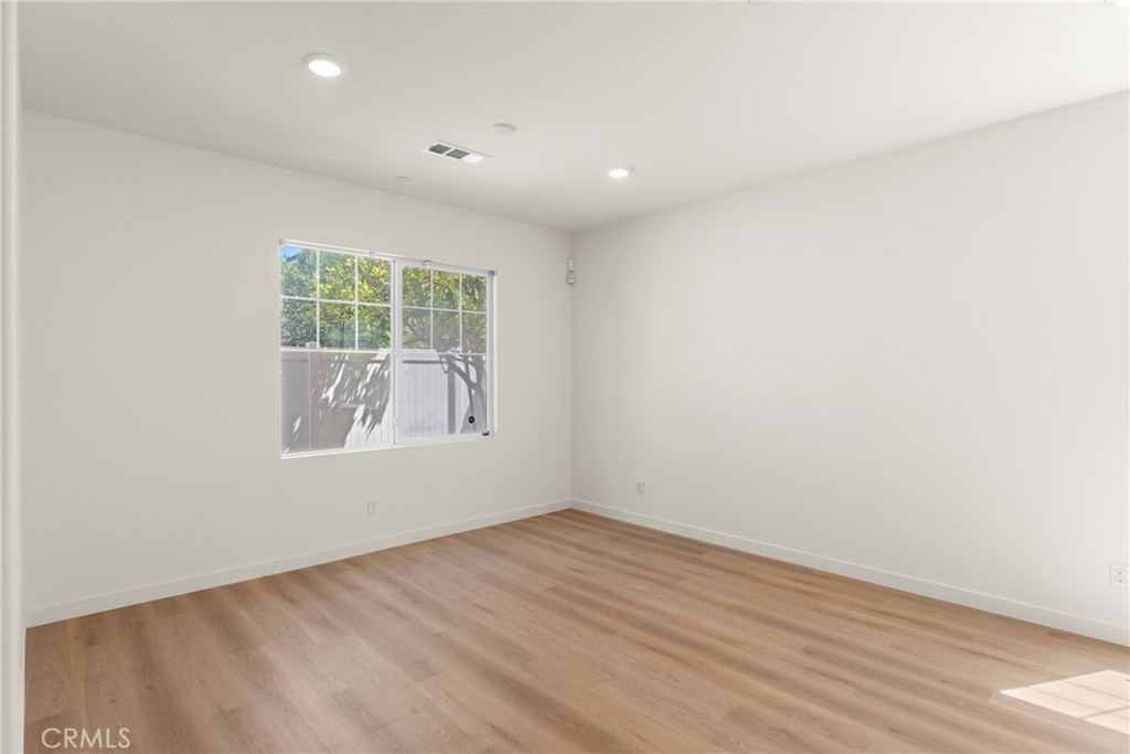 Vacant room with light wood floors, recessed lights, and a window overlooking a fenced area.