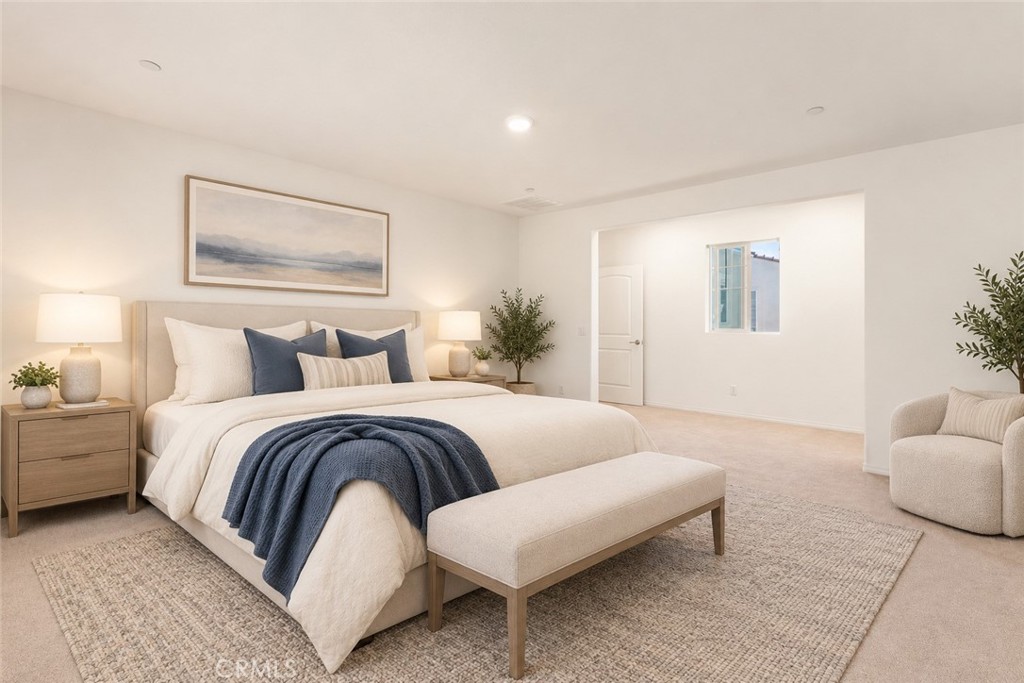 Staged bedroom with a white bed, navy throw, upholstered bench, and bright window-side sitting area.