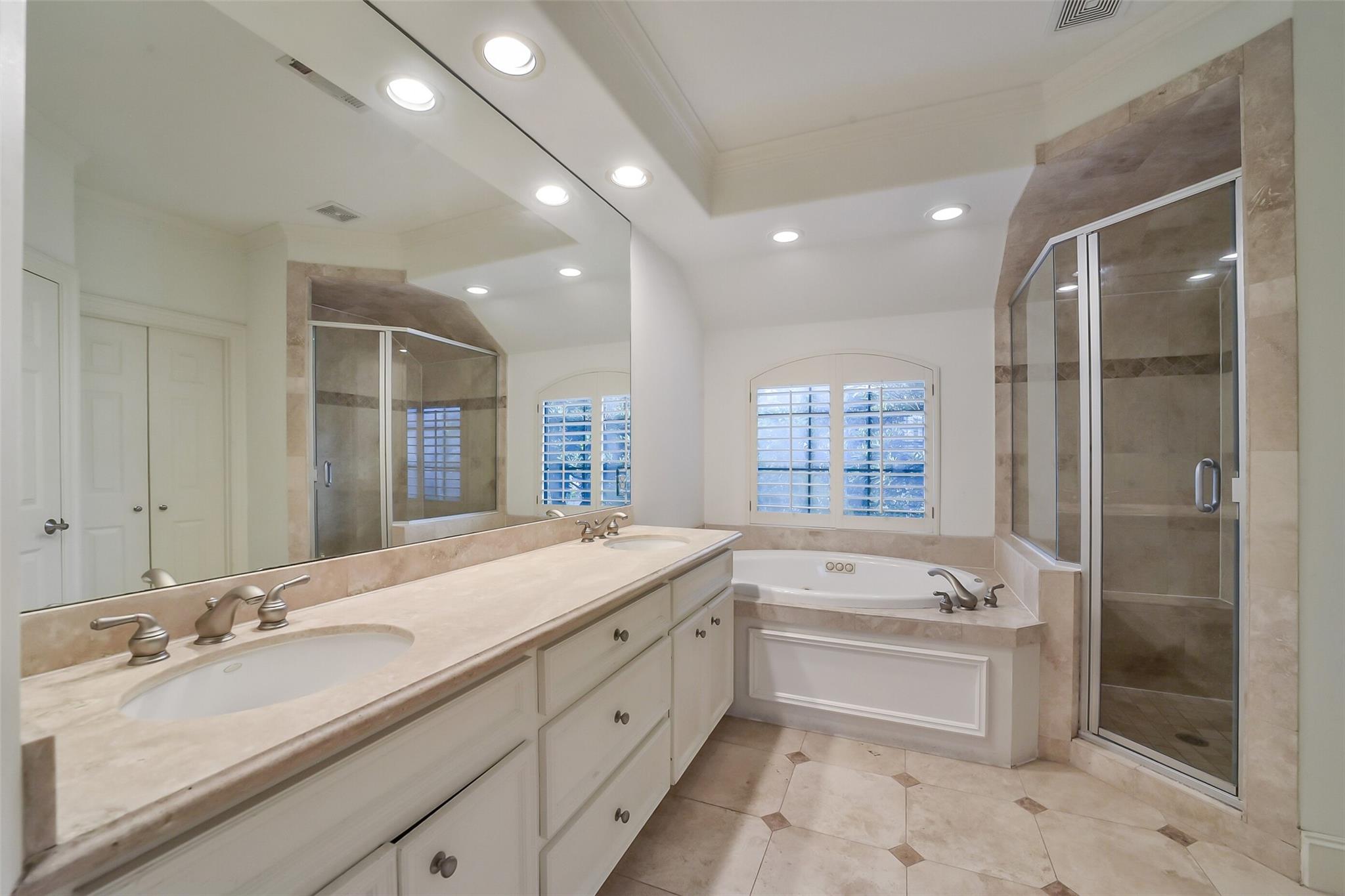 Primary bathroom with a double vanity, soaking tub beneath shuttered windows, and glass-enclosed tiled shower.