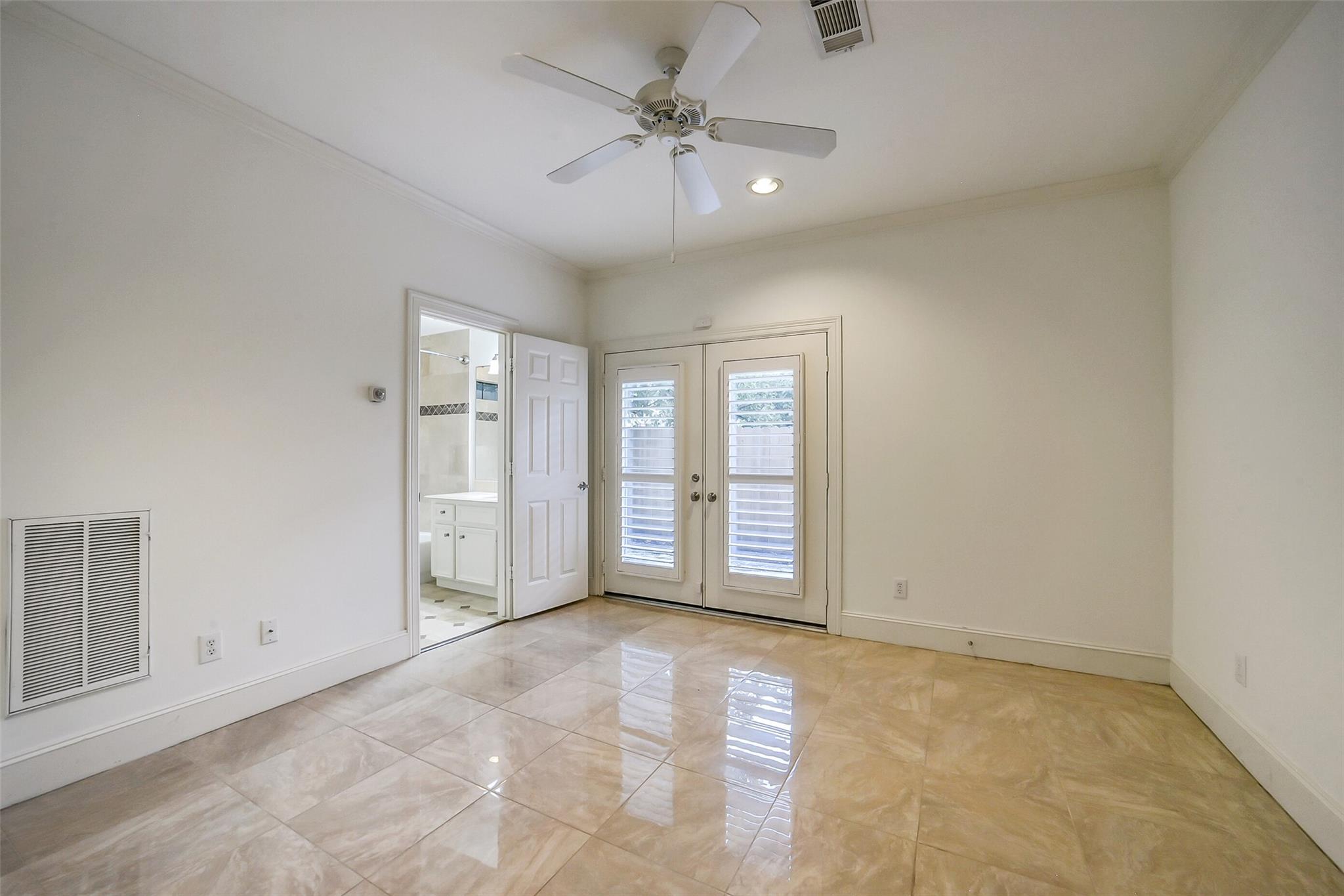 Empty tiled room with a ceiling fan, plantation-shuttered French doors, and an open bathroom doorway.