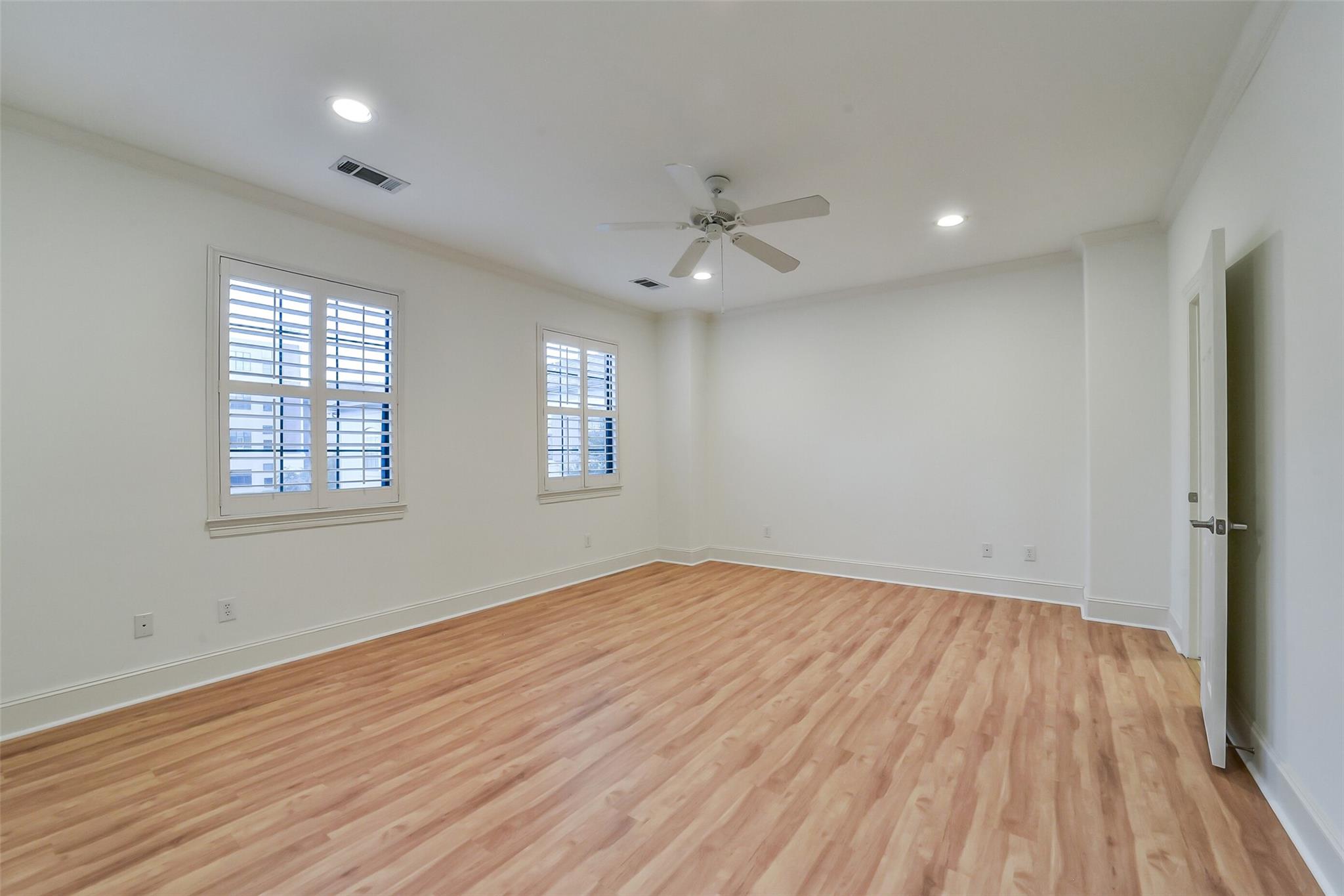 Empty room with light wood floors, two shuttered windows, and a ceiling fan.