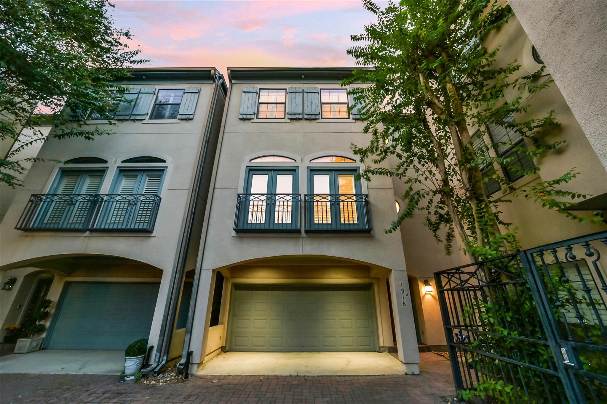 Two-story townhouse with blue-gray shutters, balcony doors, arched garage openings, and a brick-paved driveway.
