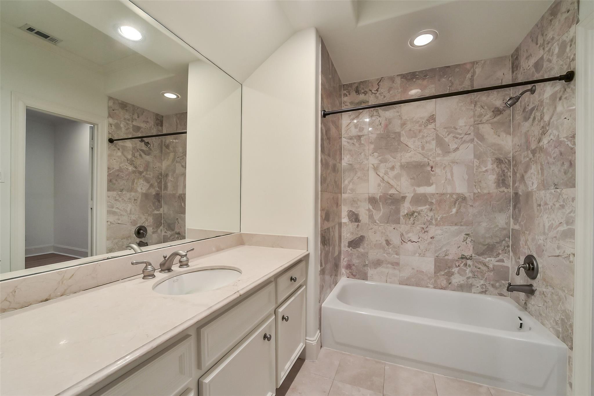 Bathroom with marble tile around a bathtub and a cream vanity with a large mirror.