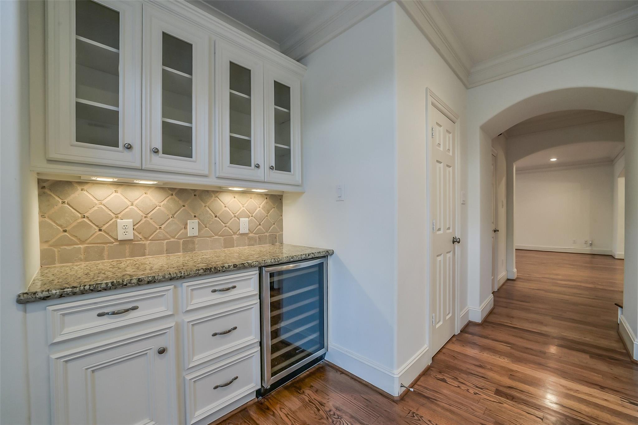 Built-in wet bar with white cabinets, stone backsplash, and wine refrigerator beside a wood-floored hallway.
