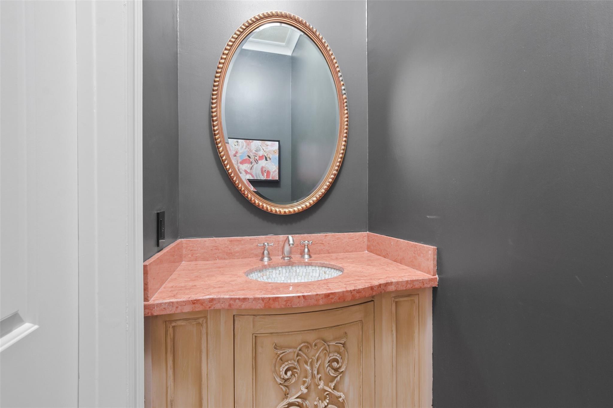 Bathroom vanity with a coral-pink stone counter, carved wood cabinetry, and ornate oval mirror.