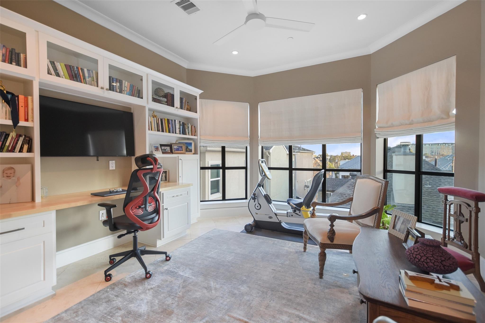 Home office with built-in bookcases, exercise bike, and large windows overlooking rooftops.
