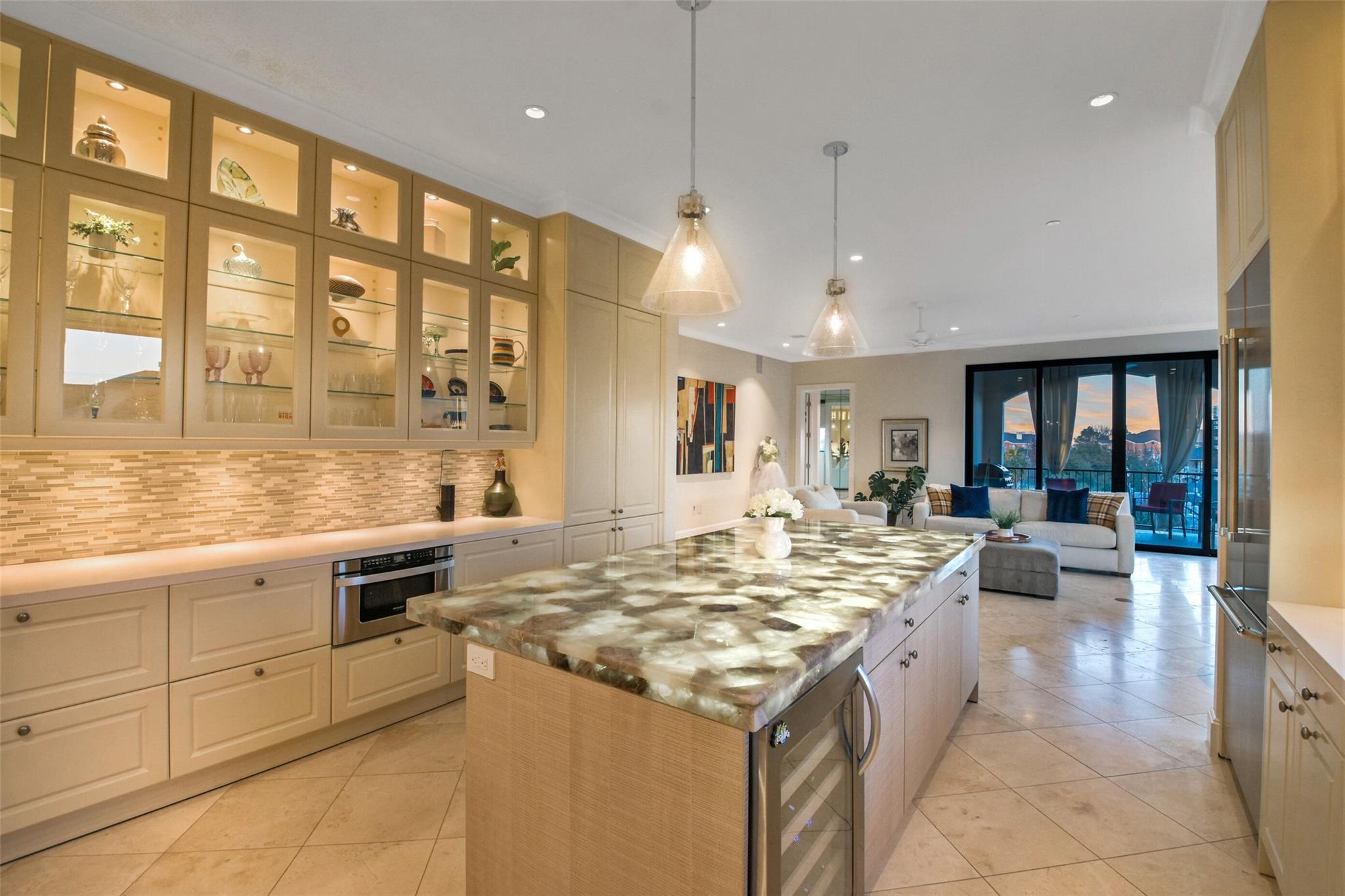 Condo kitchen with a large stone island, illuminated display cabinets, and an adjoining living room.