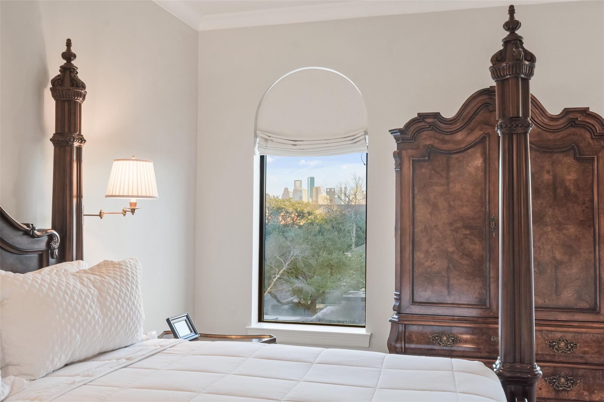 Bedroom with an ornate dark wood four-poster bed and an arched window overlooking the city skyline.
