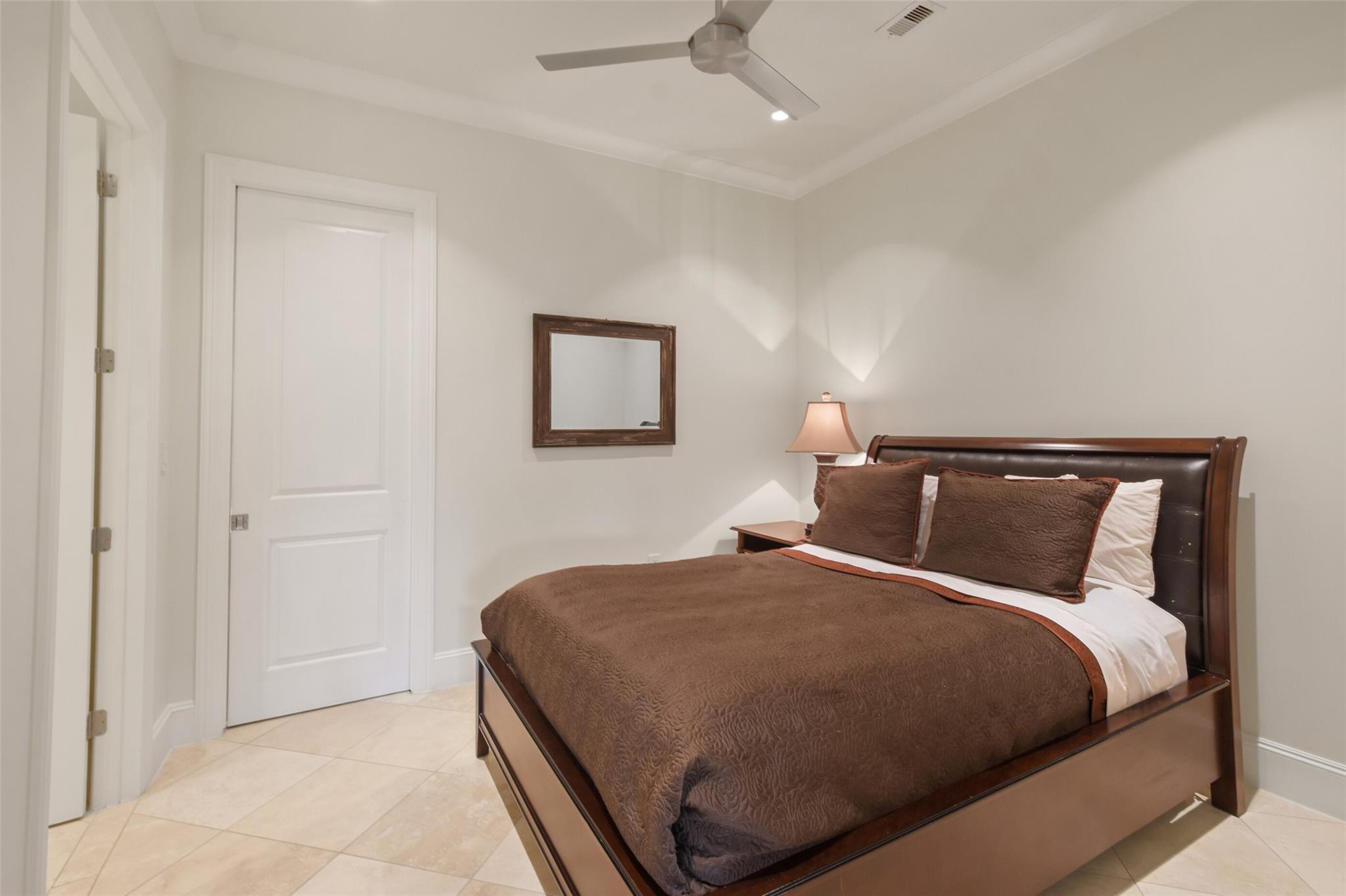 Furnished bedroom with a dark wood bed, brown bedding, pale tile floors, and a ceiling fan.