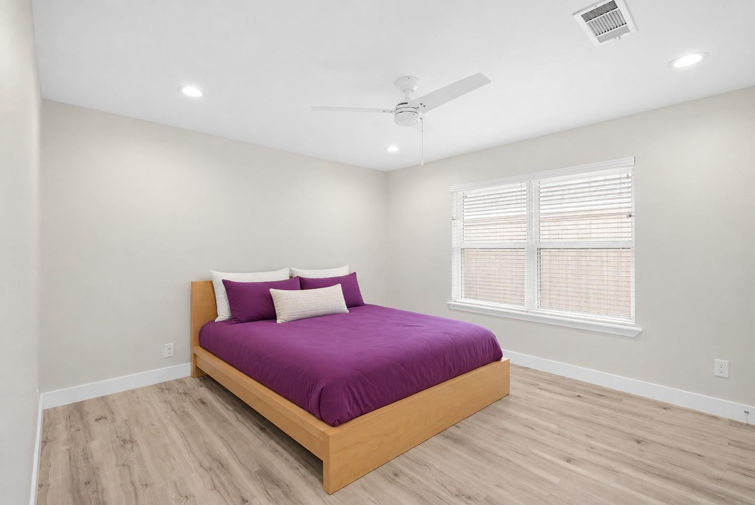 Bedroom with a low wood platform bed, purple bedding, and a wide window with white blinds.