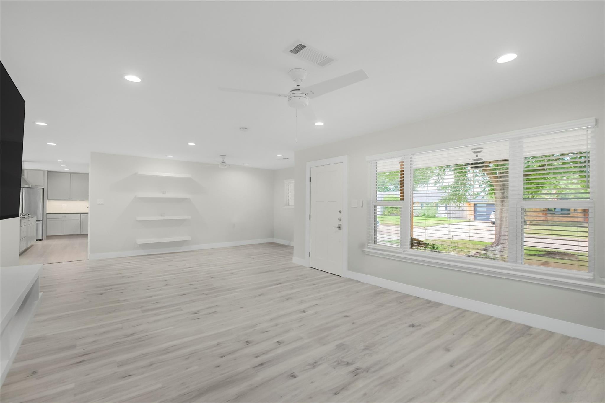 Vacant living room with pale wood-look floors, floating shelves, and wide front windows overlooking trees.
