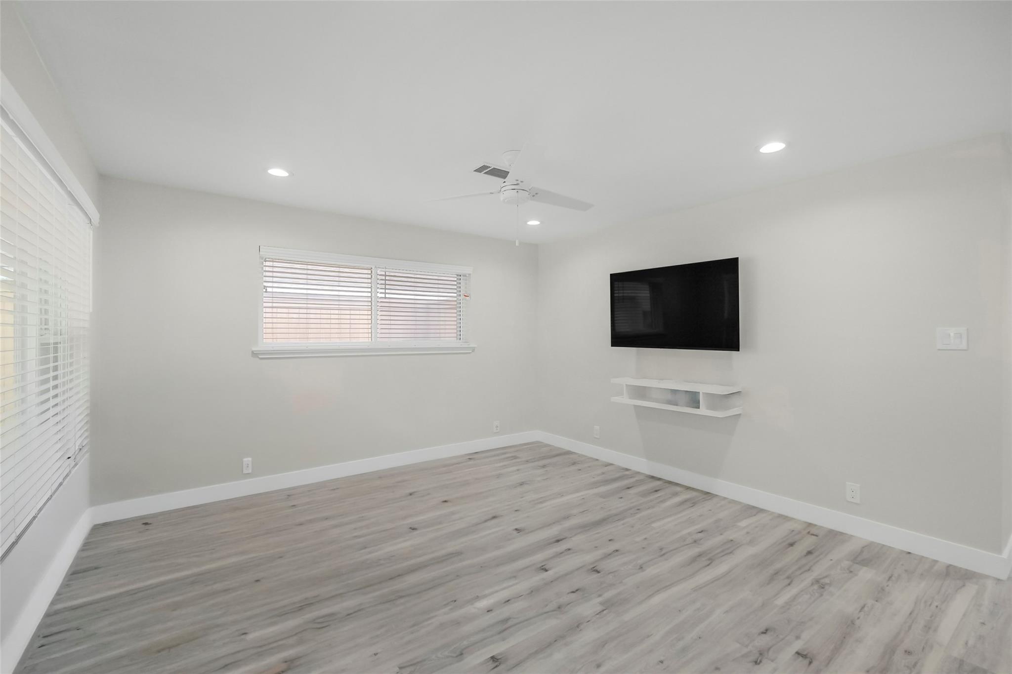 Bright empty room with gray plank flooring, window blinds, and a wall-mounted television.
