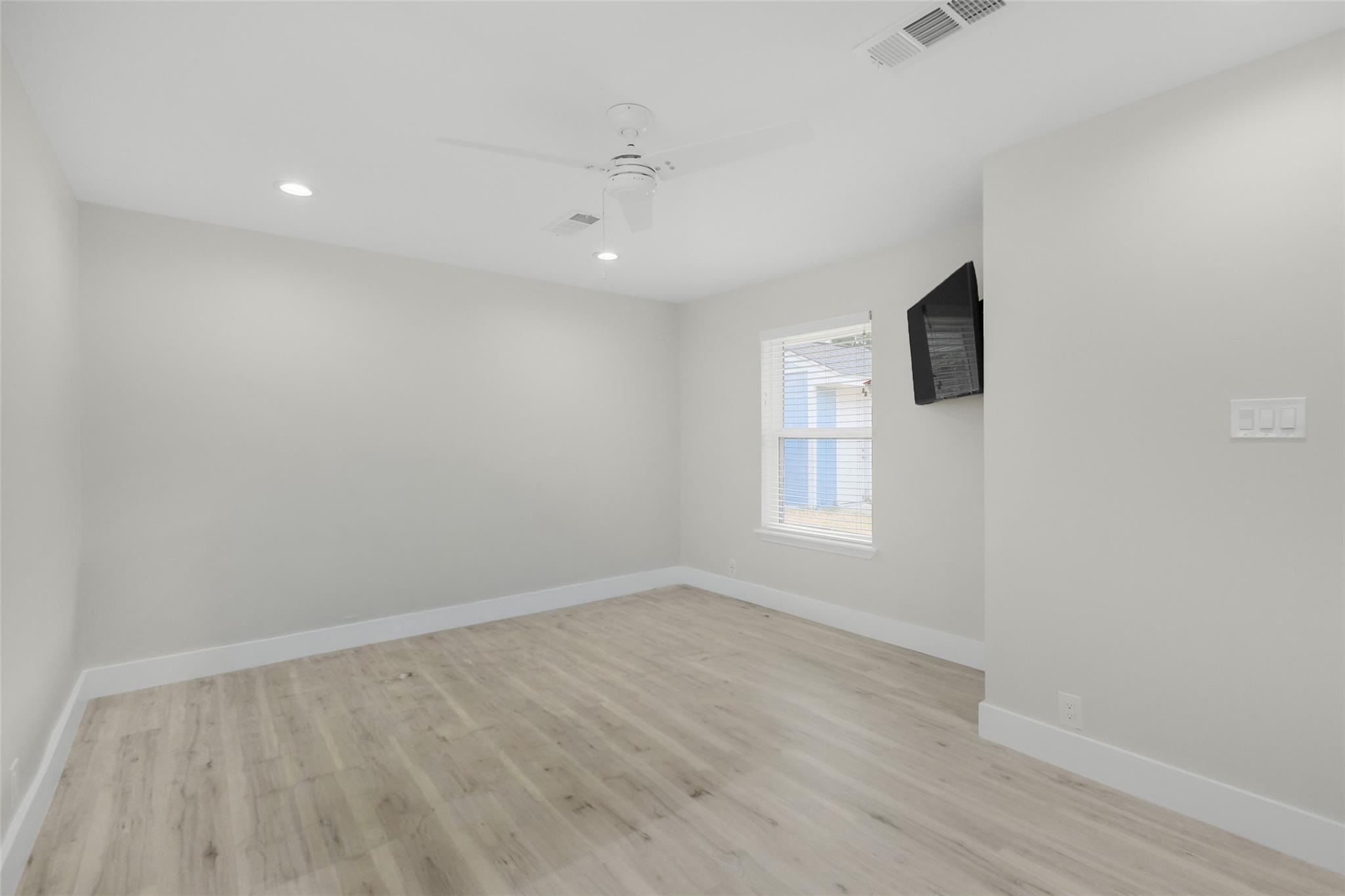 Empty bedroom with pale wood flooring, a window with blinds, and a wall-mounted television.