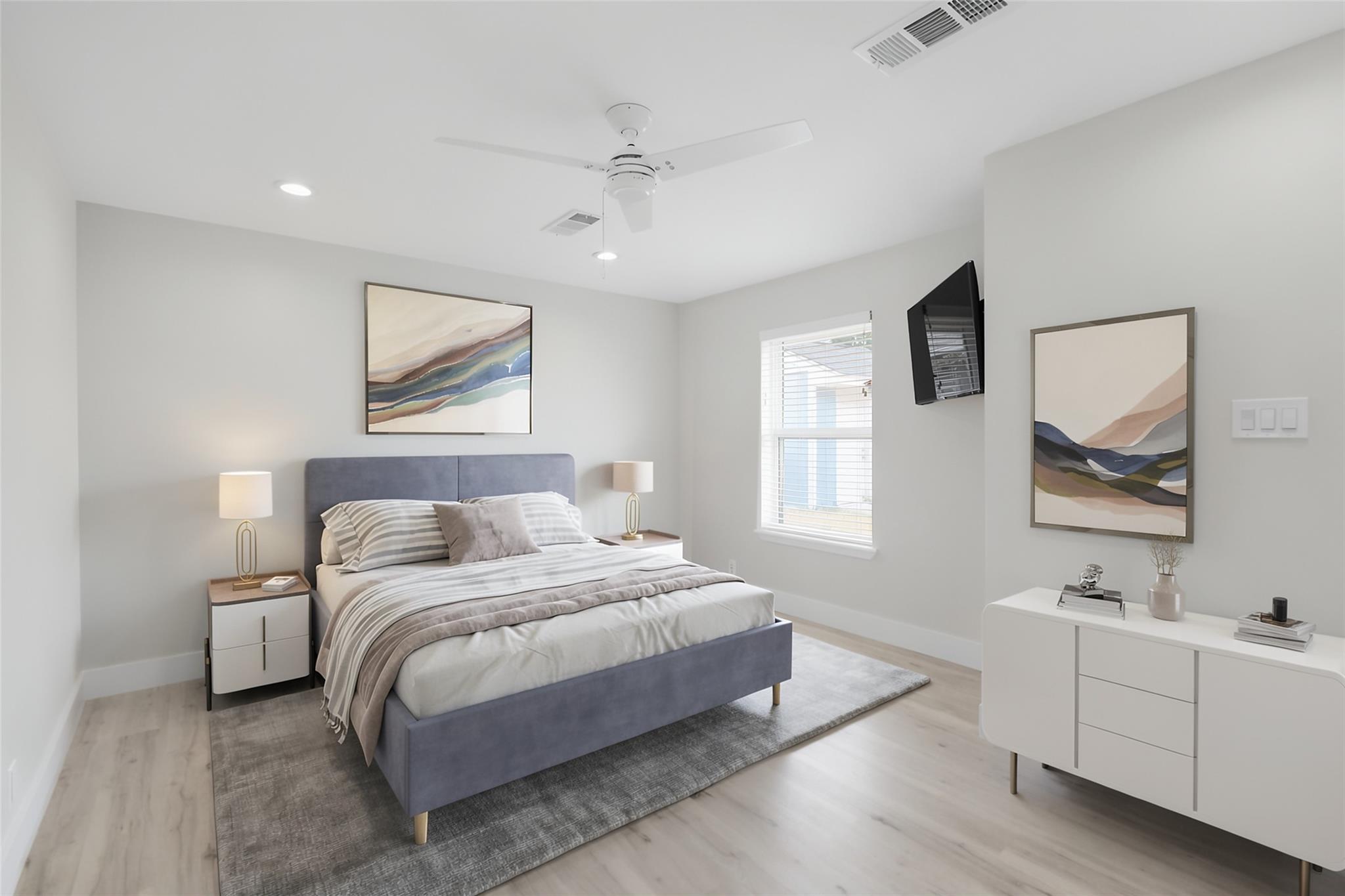 Staged bedroom with a blue-gray bed, striped bedding, a window with blinds, and a wall-mounted television.