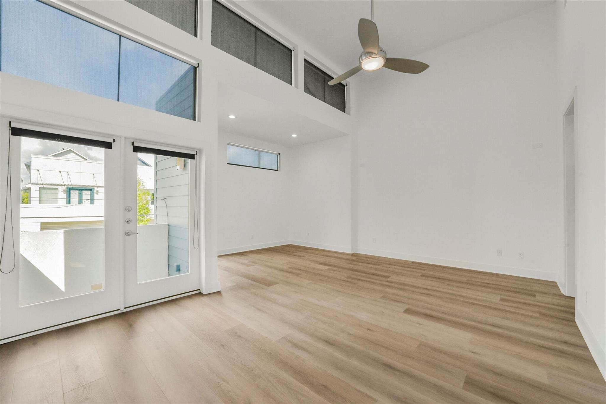 Empty living room with high ceiling, light wood floors, clerestory windows, and glass balcony doors.