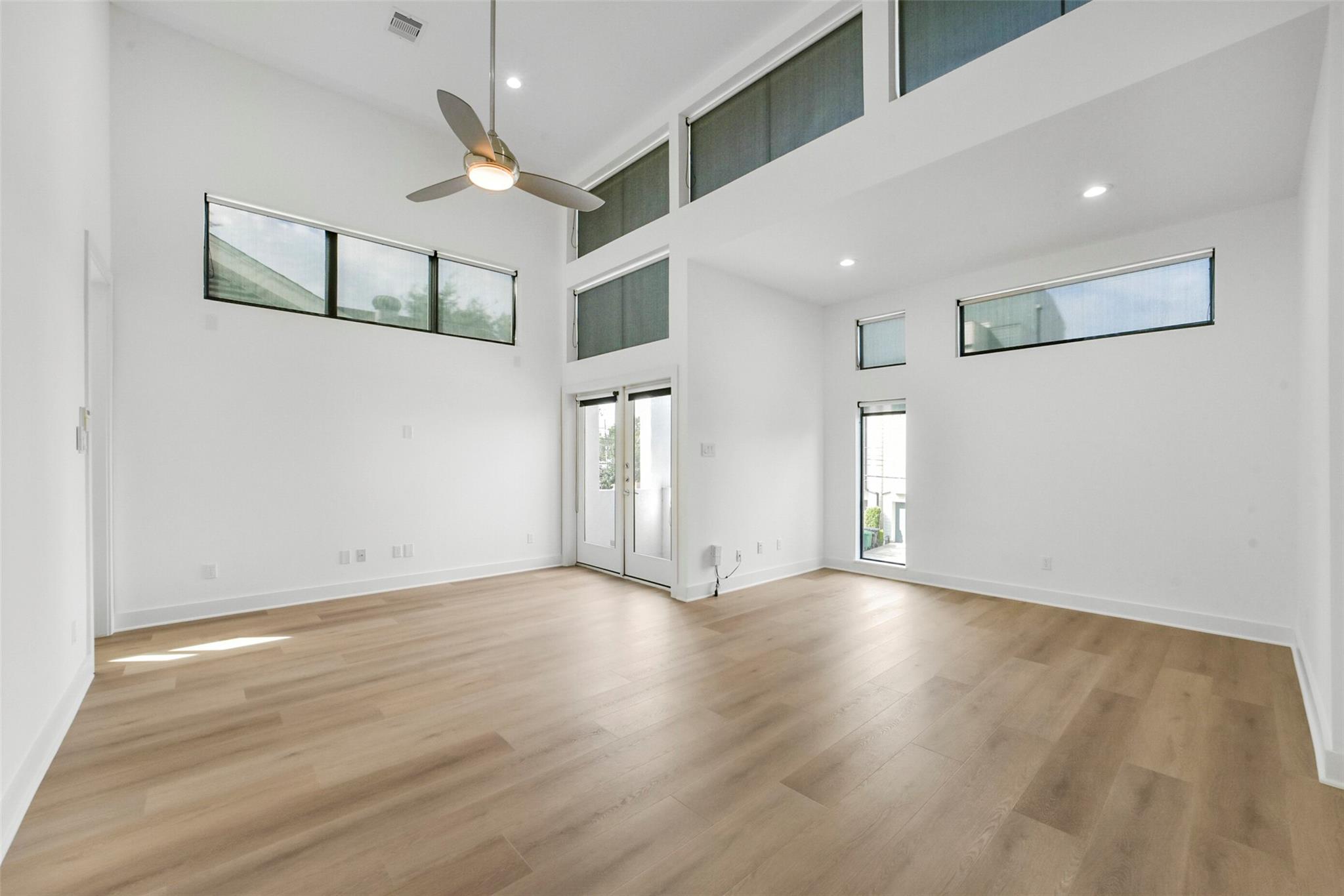 Vacant double-height living room with light wood floors, tall windows, and a ceiling fan.