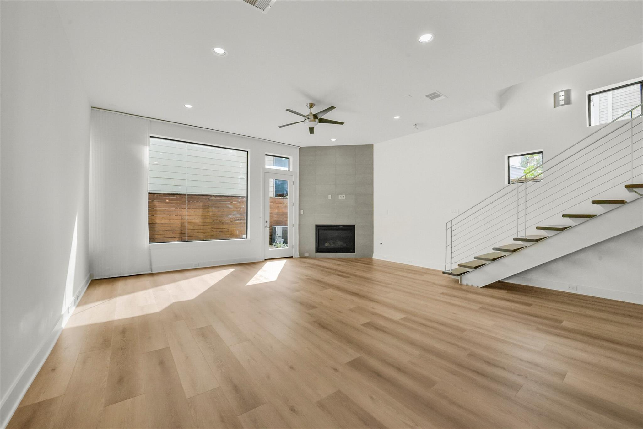 Vacant living room with light wood floors, a tiled fireplace, large windows, and an open staircase.