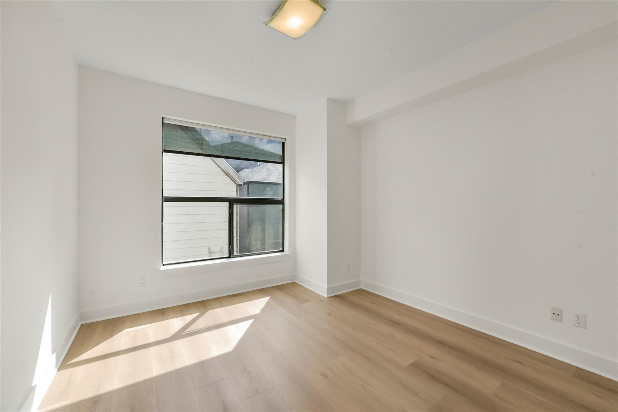 Empty bedroom with light wood floors and sunlight streaming through a large window.