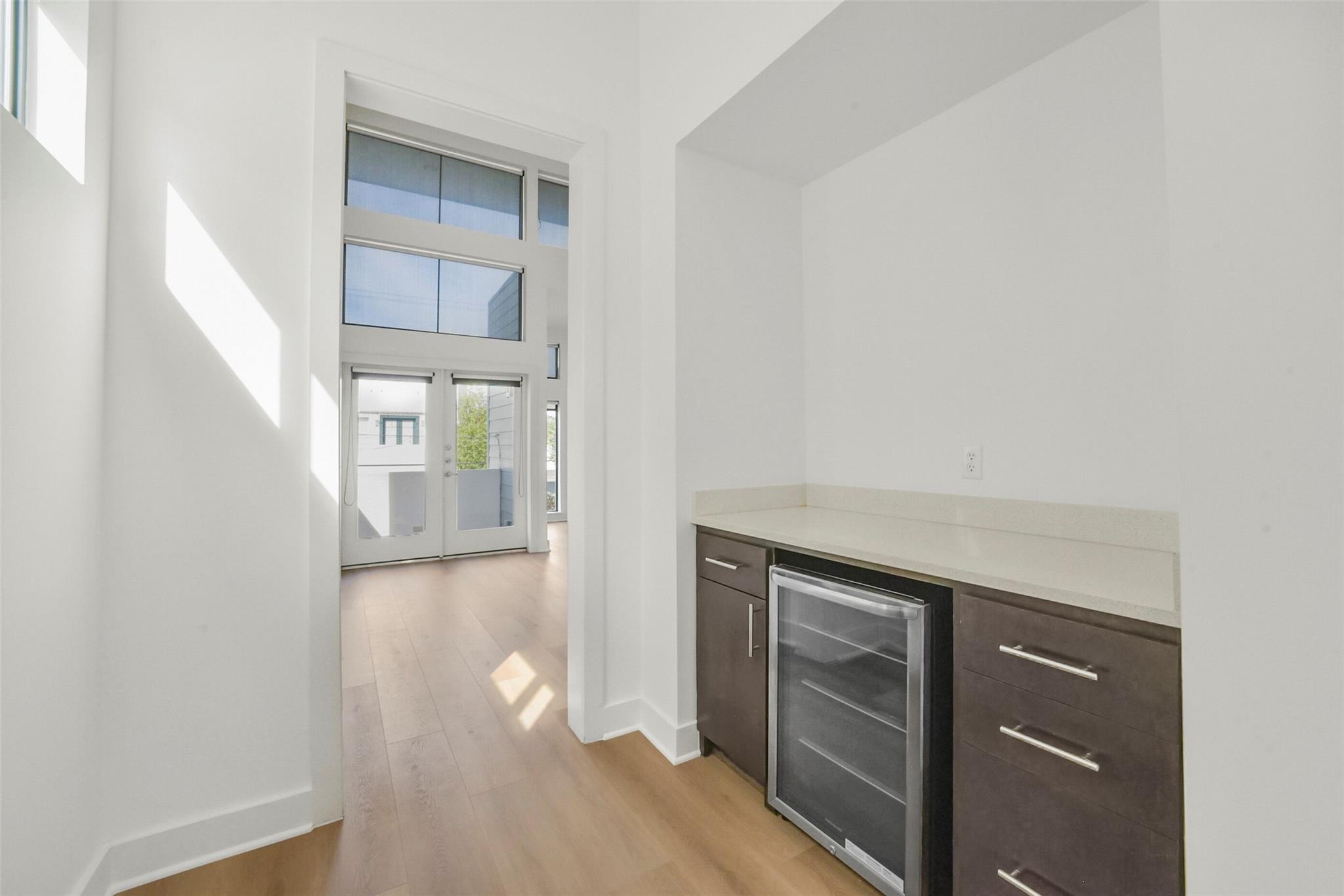 Built-in beverage bar with a glass-front wine refrigerator beside a sunlit room with tall windows.