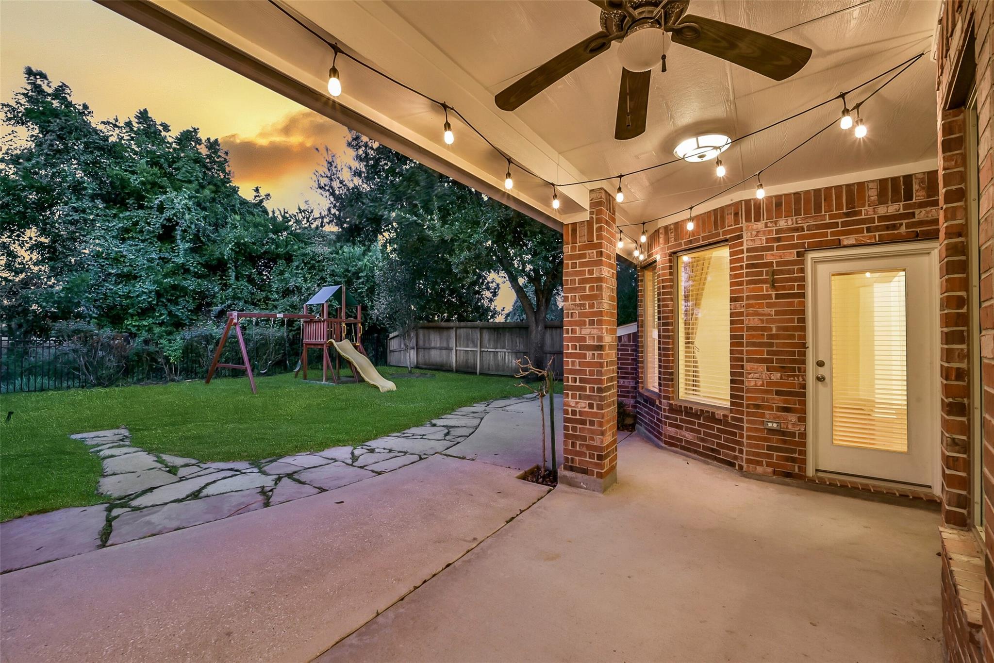 Covered brick patio with string lights and a ceiling fan overlooking a lawn and children's playset at dusk.