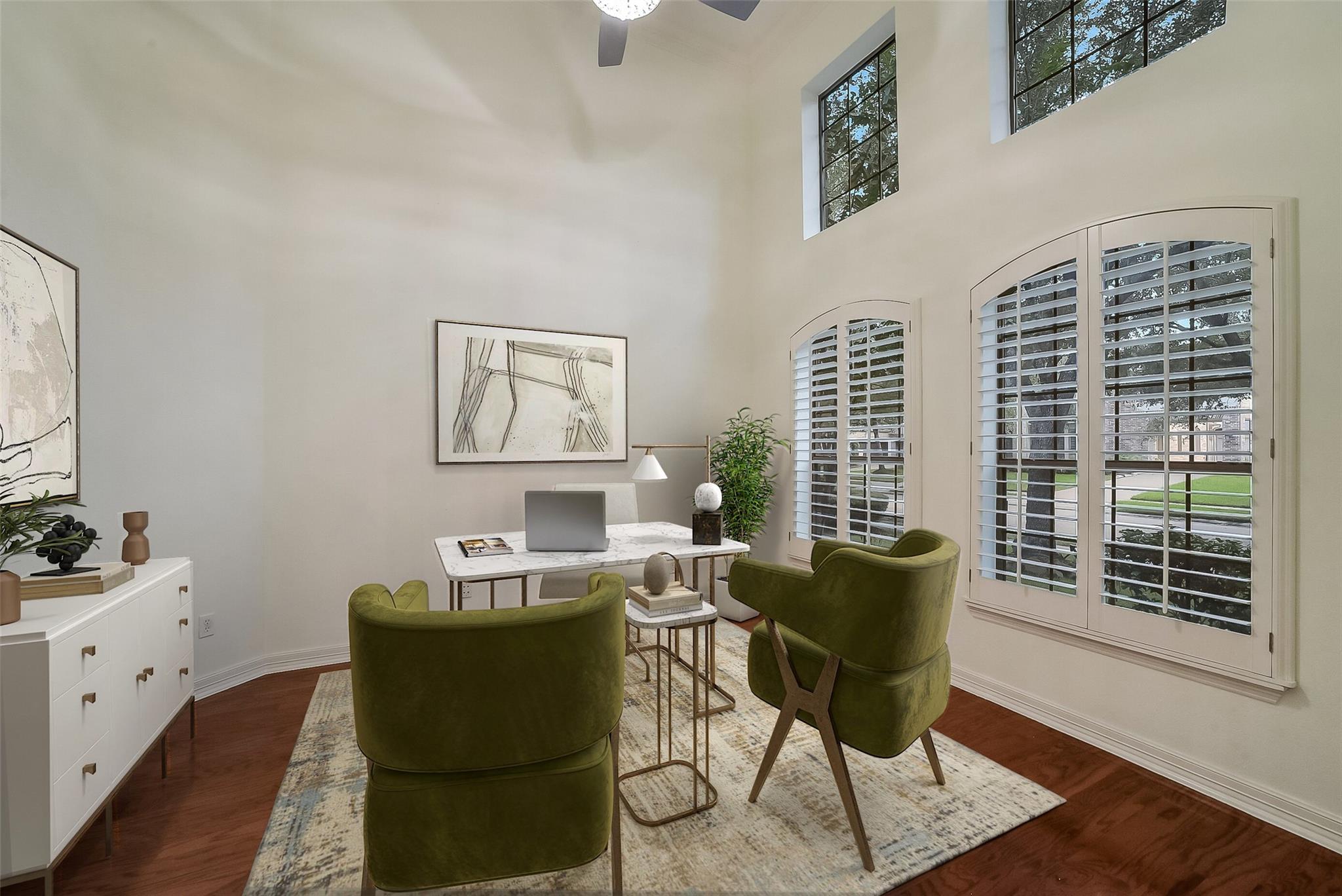 Home office with olive-green armchairs, a desk, plantation shutters, and a high ceiling.