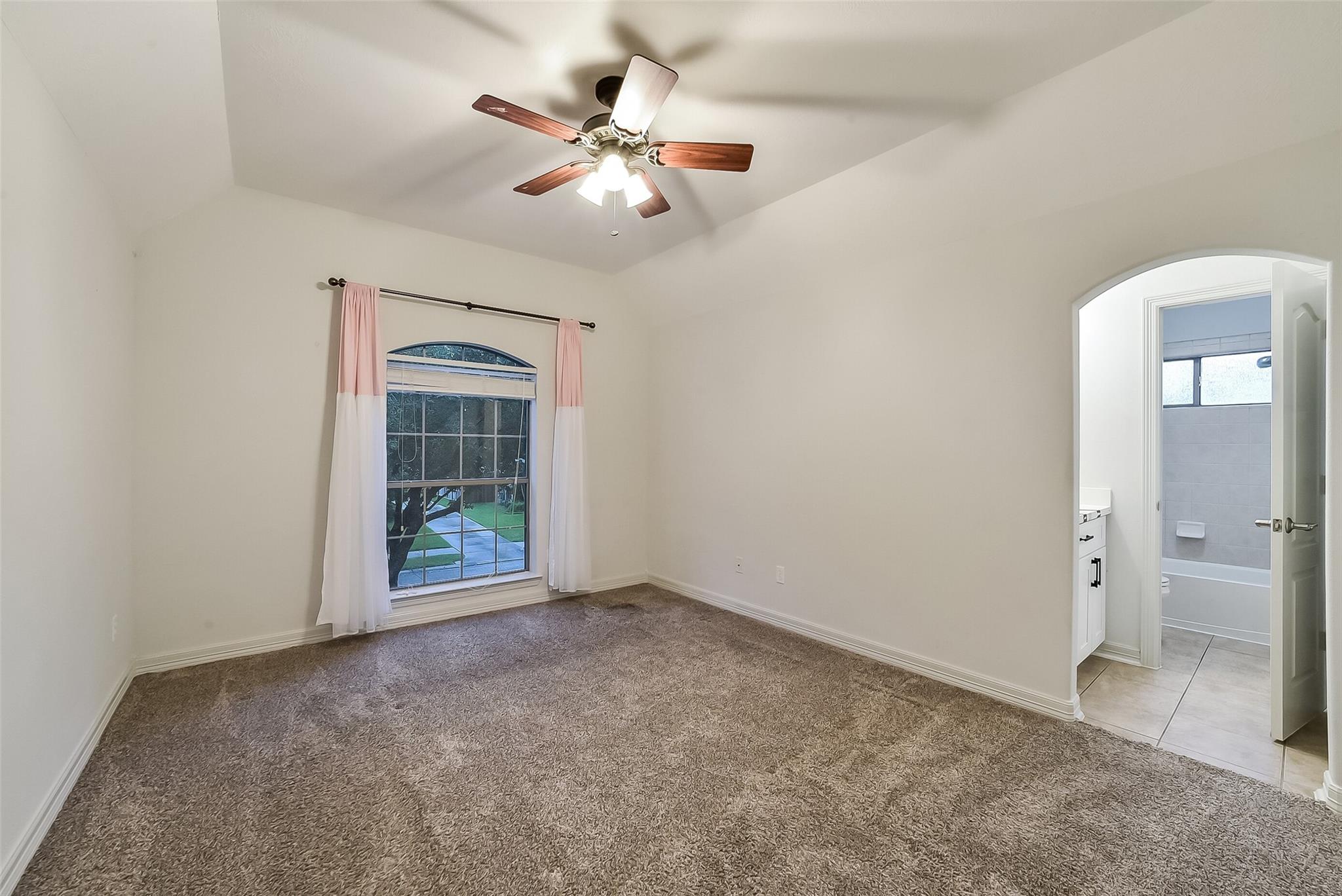 Empty carpeted bedroom with a ceiling fan, curtained window, and doorway to an adjoining bathroom.