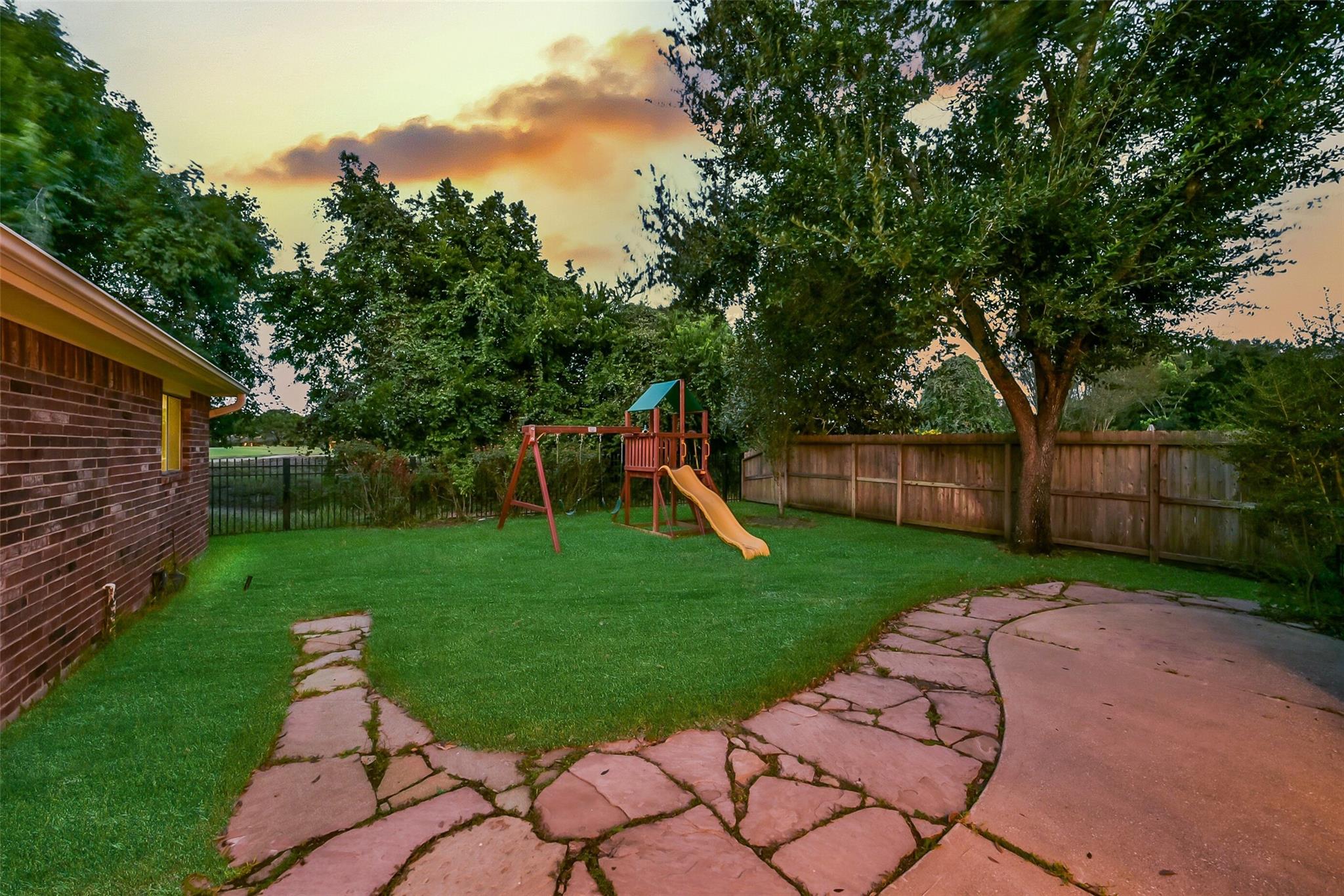 Backyard with a children's playset, mature trees, fenced lawn, and curved flagstone patio.